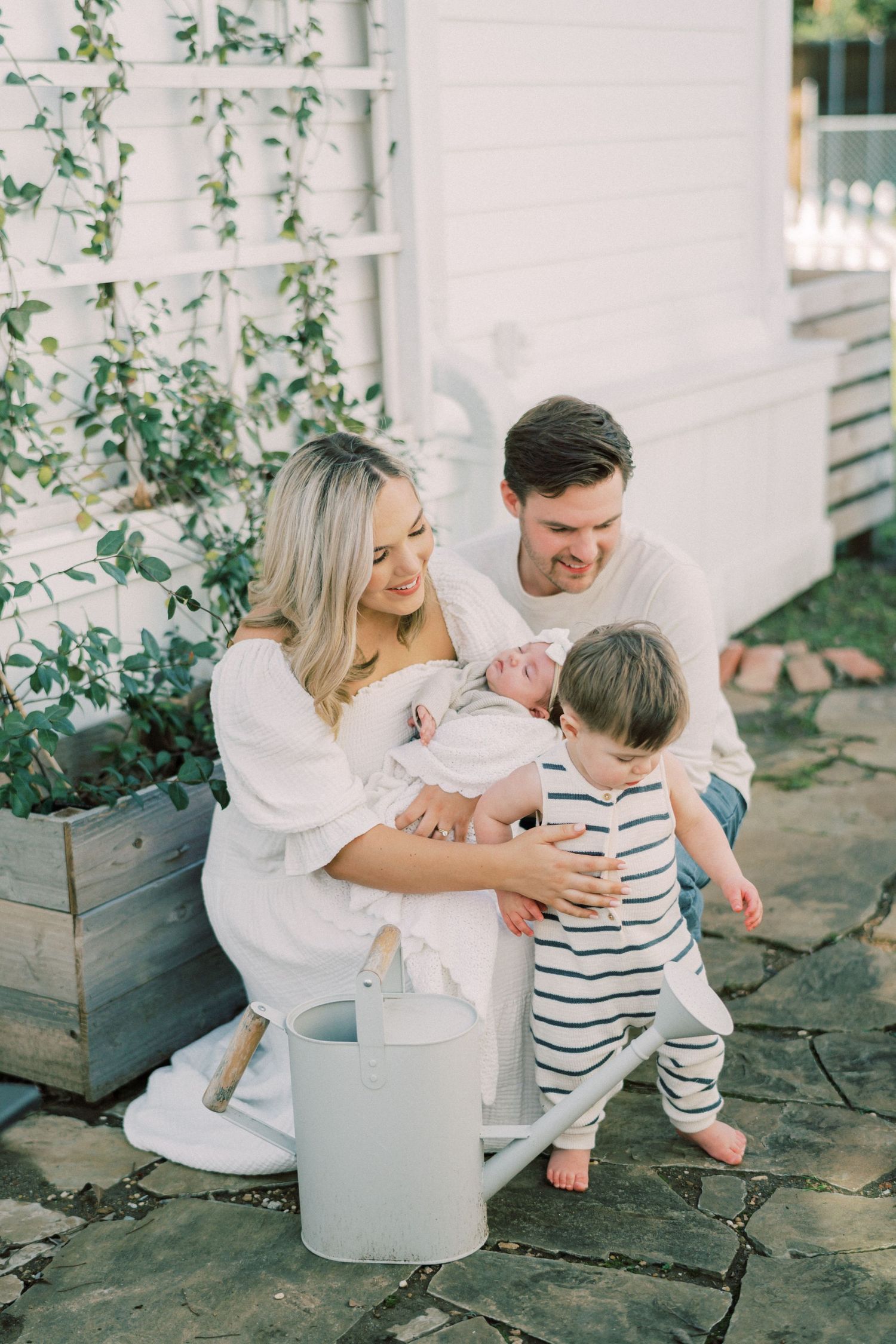 Family in white clothing sits together on stone patio with climbing vines and watering can in rustic garden setting.