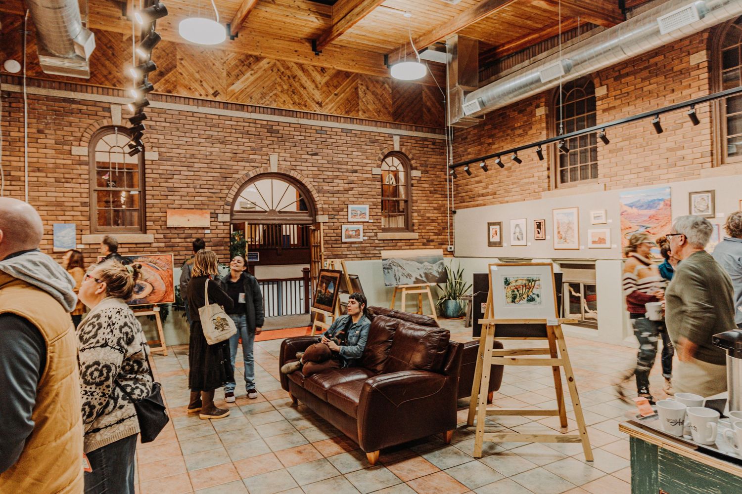 Art gallery space with brick walls, vintage furniture, and people browsing artwork.