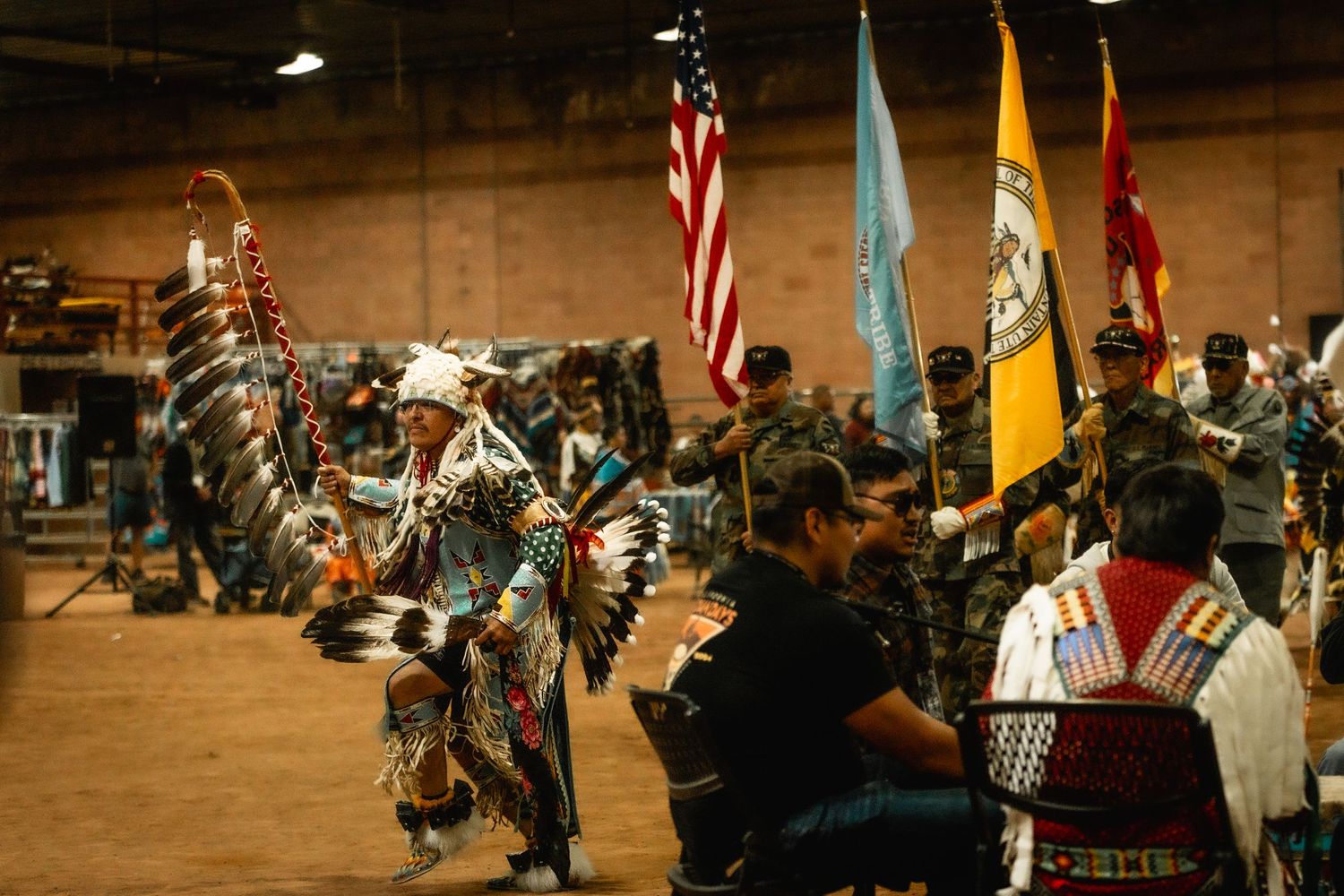 A pow wow celebration features American flags and traditional dancers performing in an indoor arena.