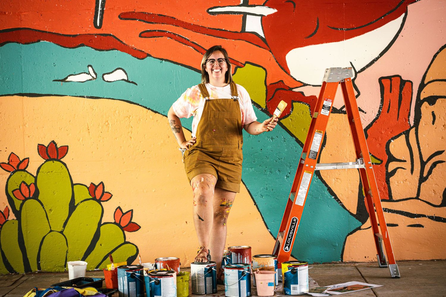Artist in overalls stands on ladder in front of colorful desert mural with paint cans below.