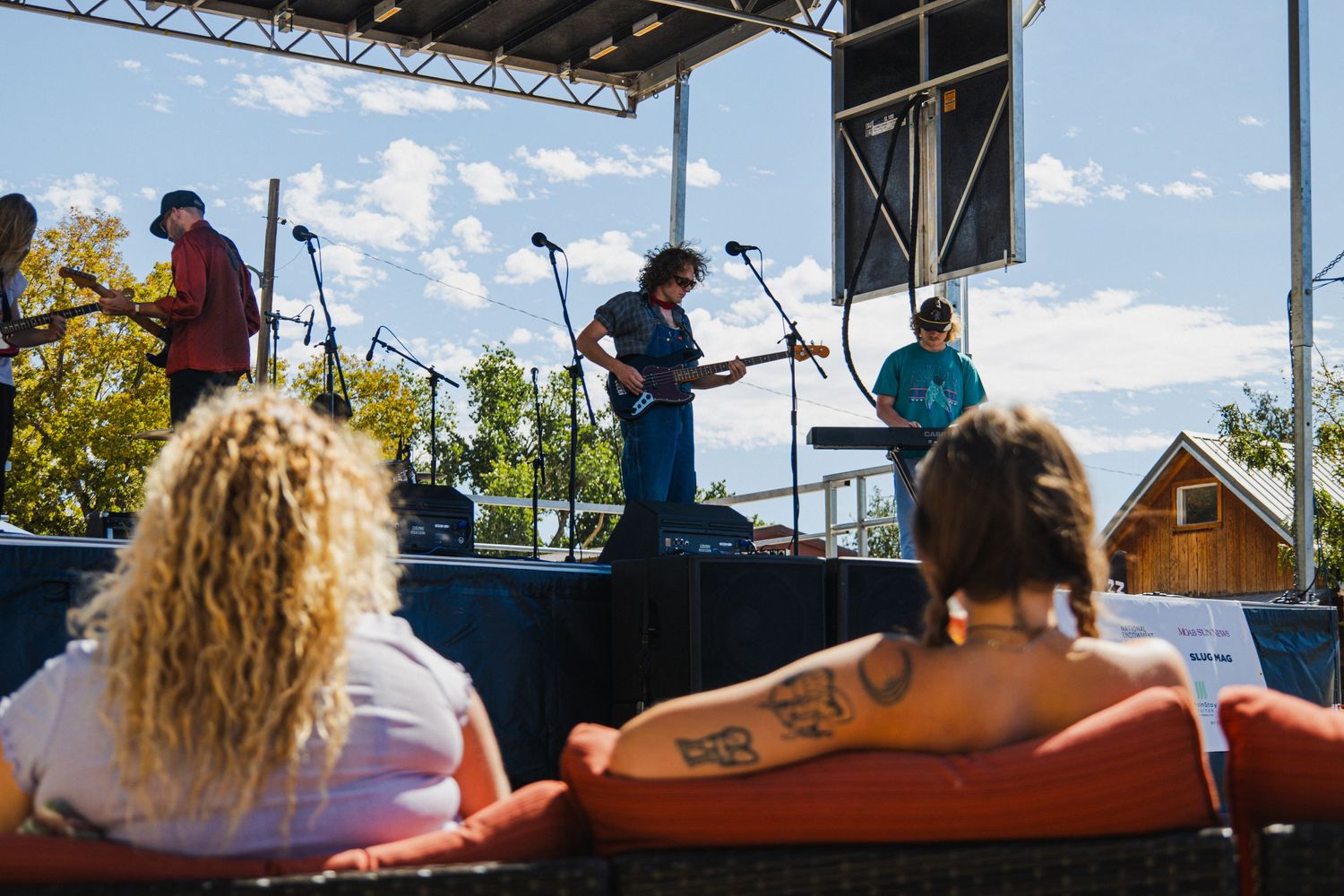 Outdoor festival stage setup with musical equipment beneath a metal framework structure against a bright blue sky.