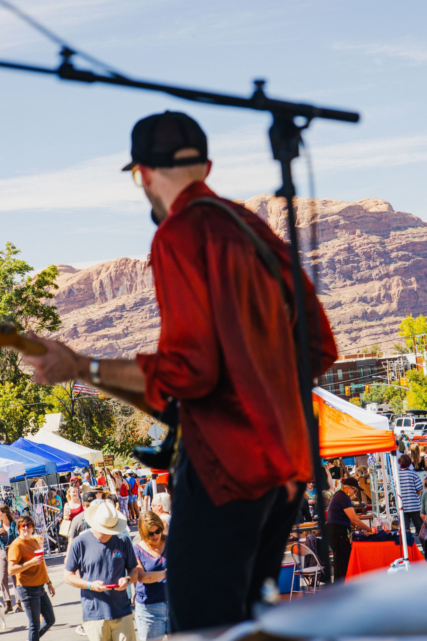 Musician performs on stage against backdrop of red rock cliffs at outdoor market festival.