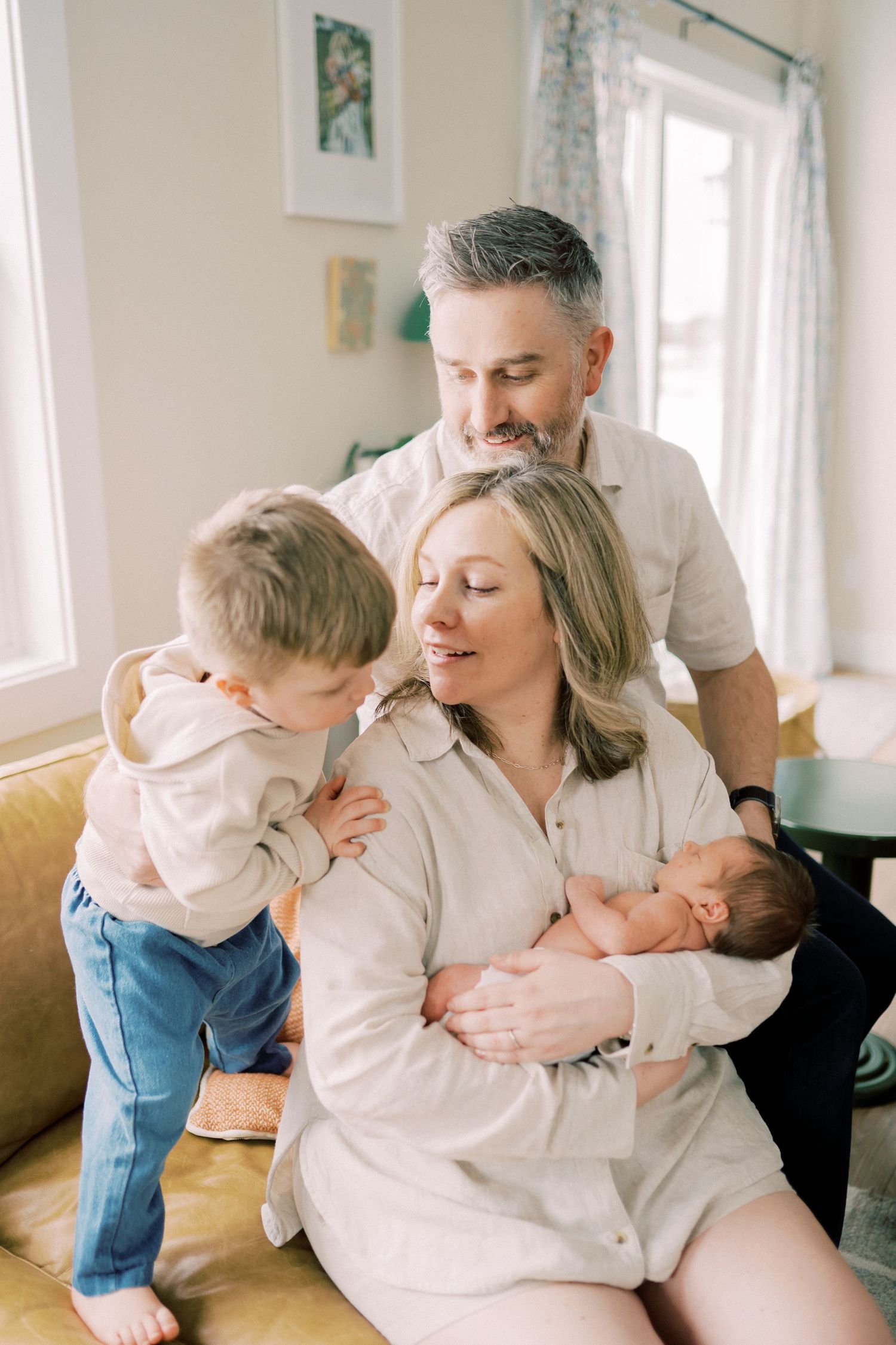 A family sits together in a sunlit living room while caring for a small baby and toddler.