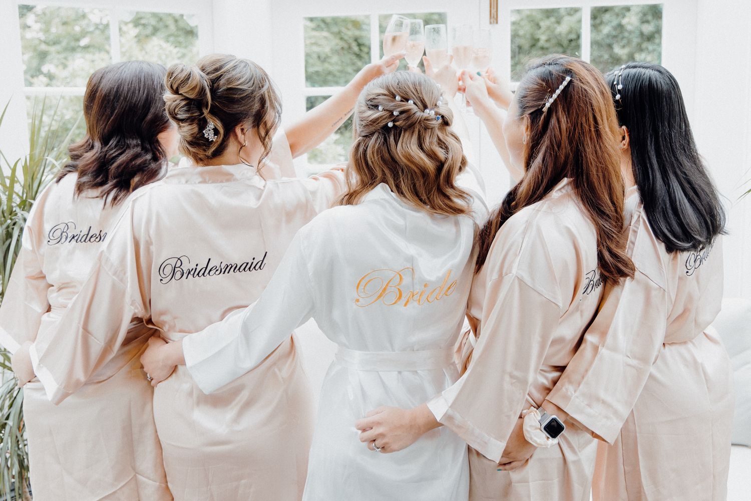 Group of bridesmaids wearing matching light pink satin robes with gold lettering captured by Apertured Media Auckland Wedding Photographer.