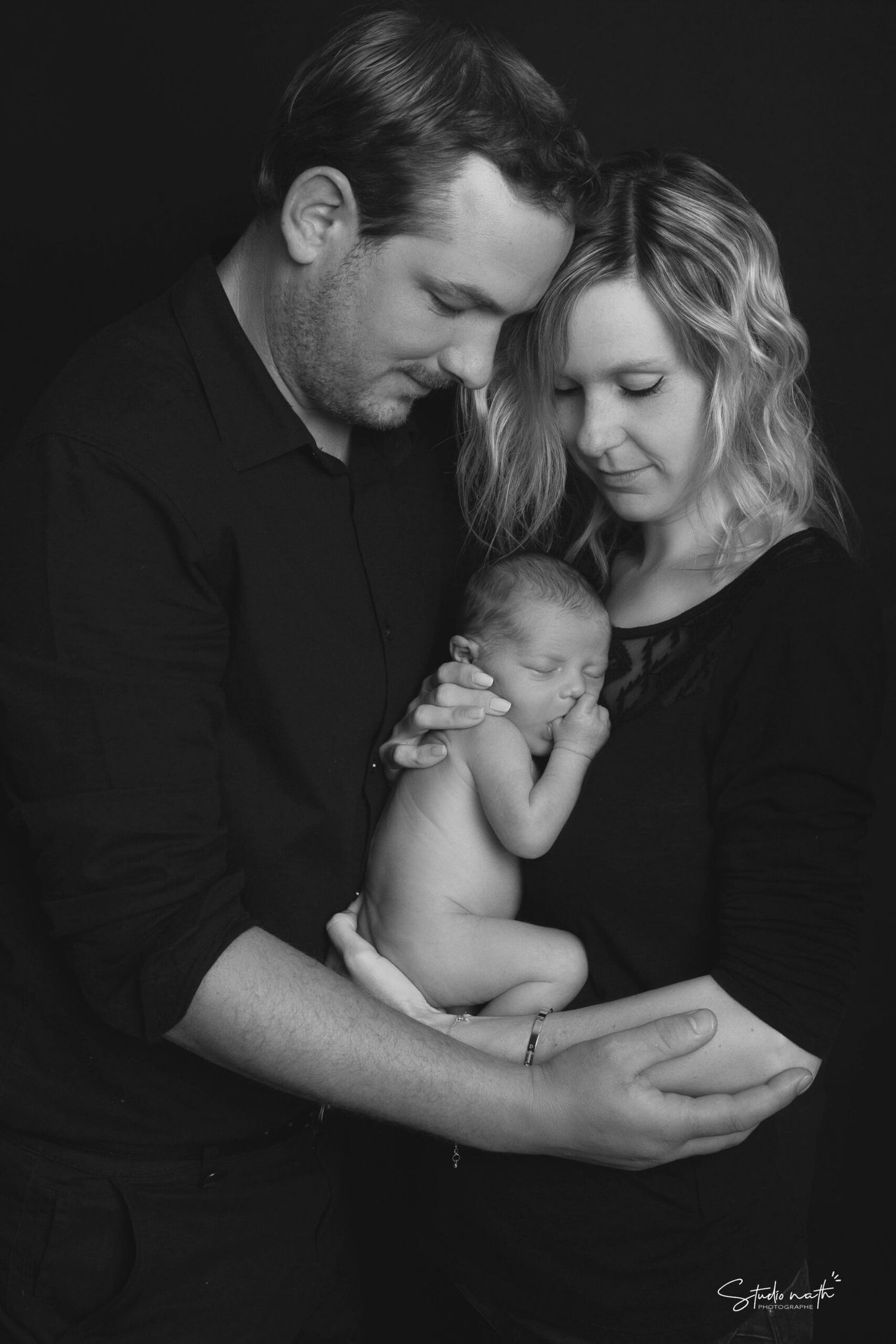 A tender black and white studio portrait of parents cradling their newborn baby against a dark background.