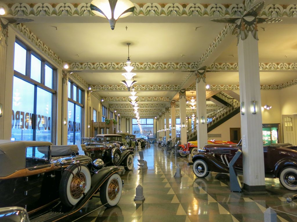 Vintage automobiles line the showroom floor of an elegant automotive museum with ornate ceiling details and large windows.