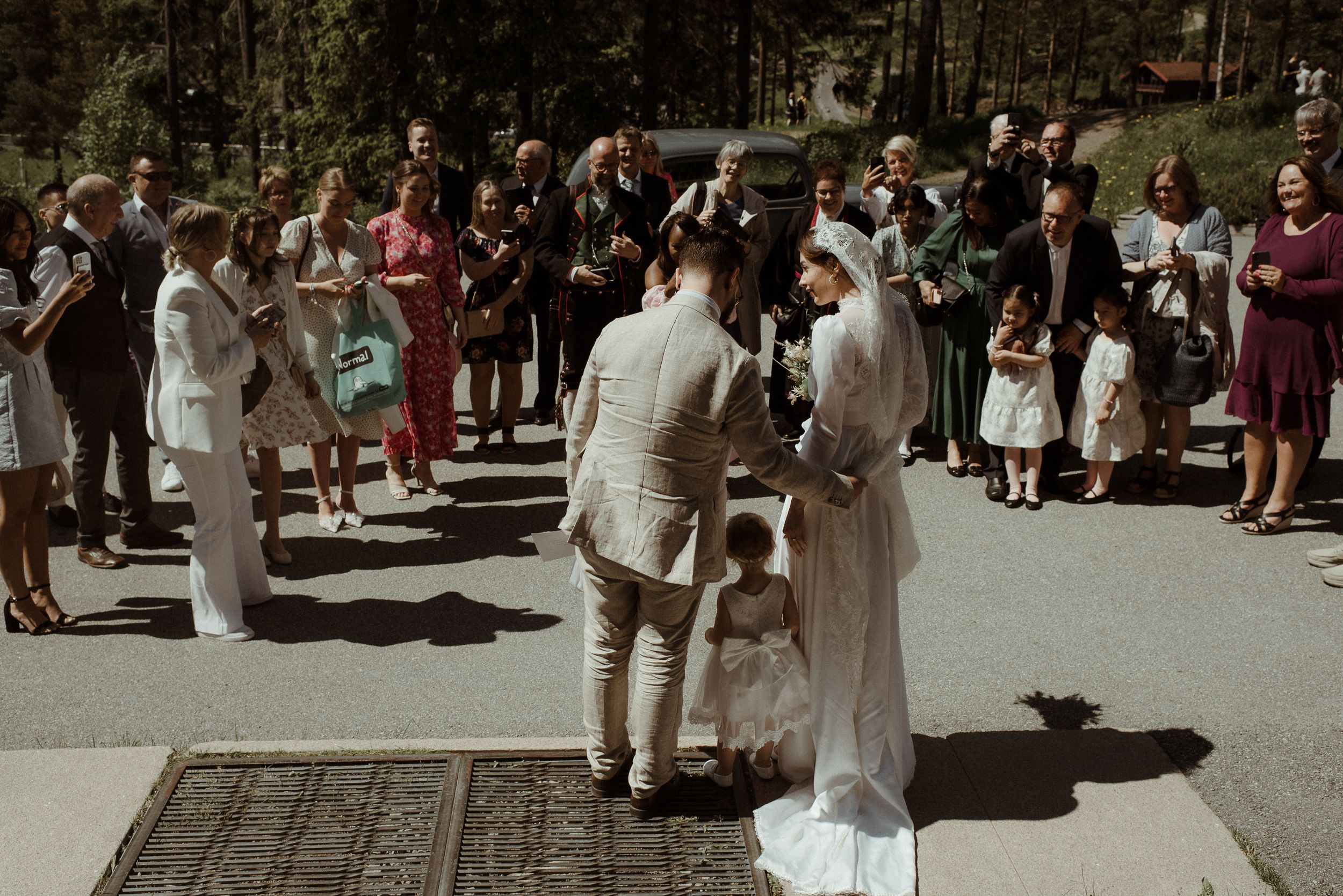 Wedding guests gather outside in sunlight on a paved walkway surrounded by trees.