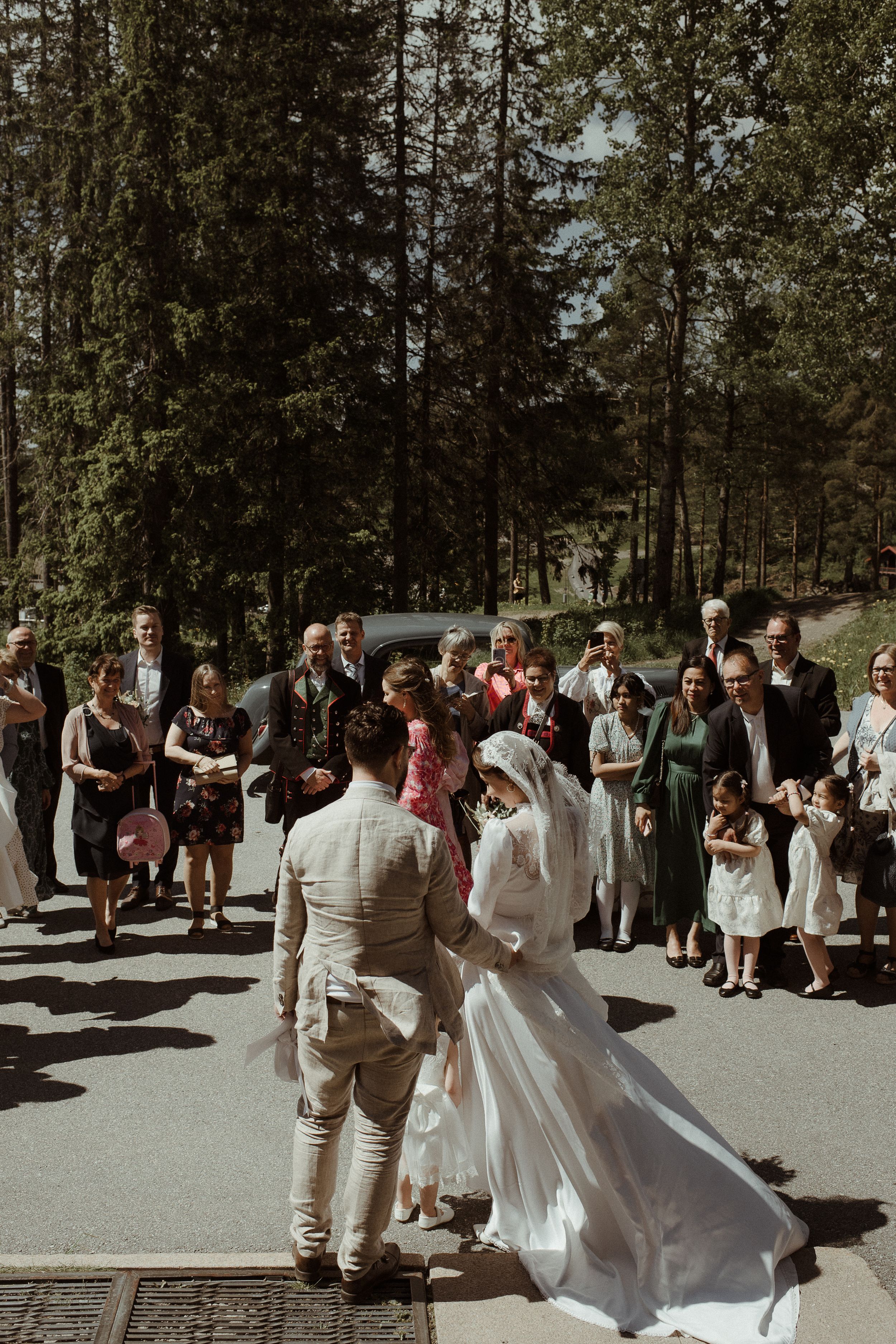 A wedding celebration outdoors with guests gathered in a wooded setting on a sunny day.
