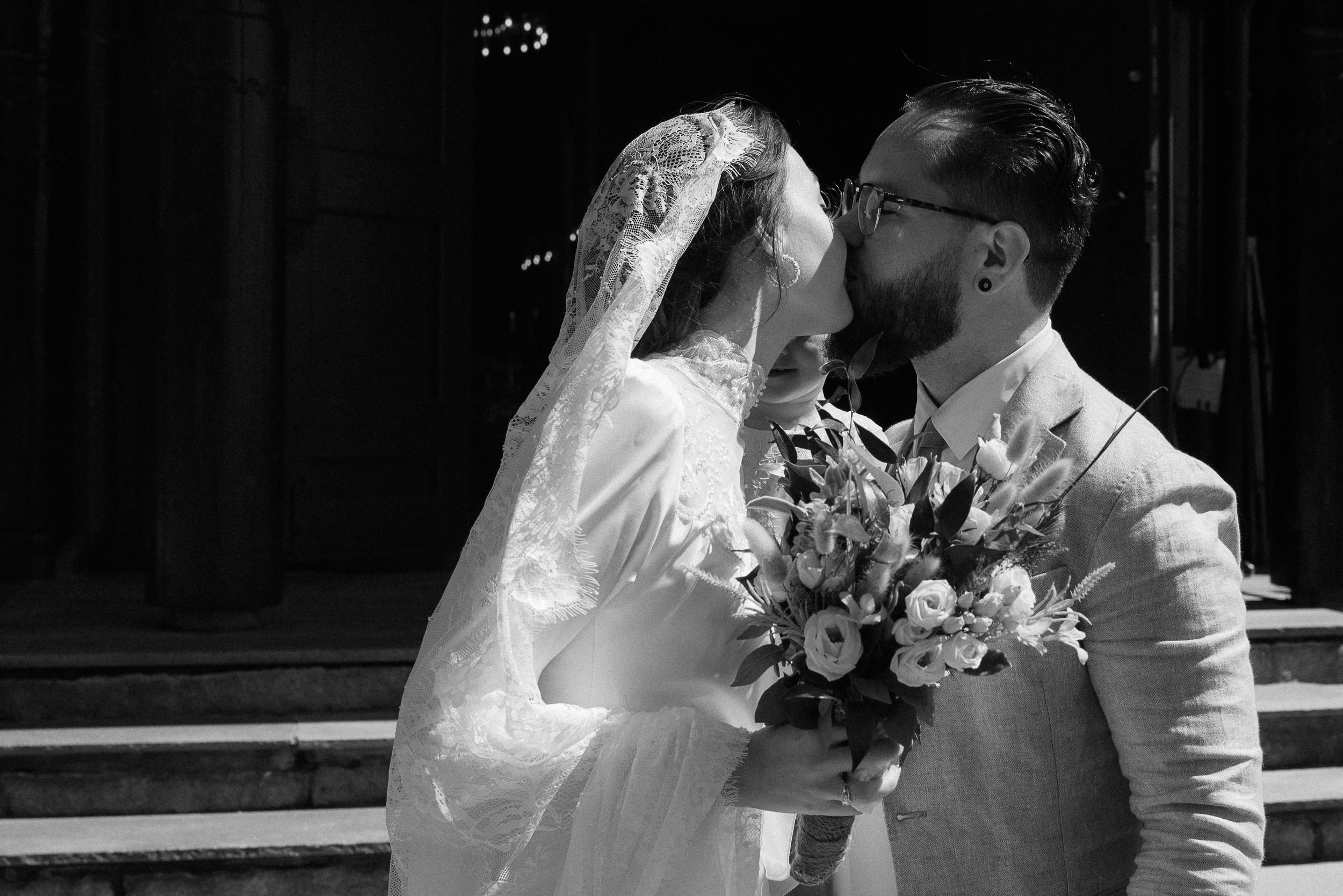 A bride and groom share a romantic kiss outside a church while holding a floral bouquet.