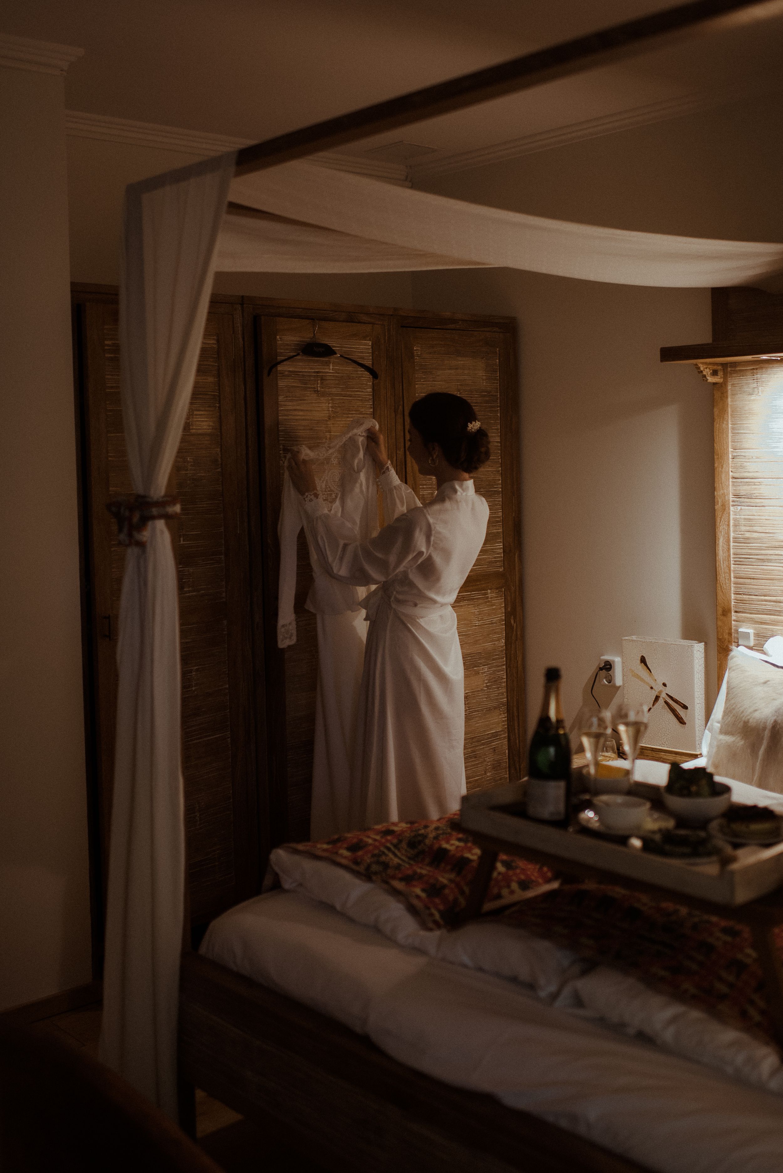 White wedding dress hanging on wooden wardrobe door in moody bedroom lighting.