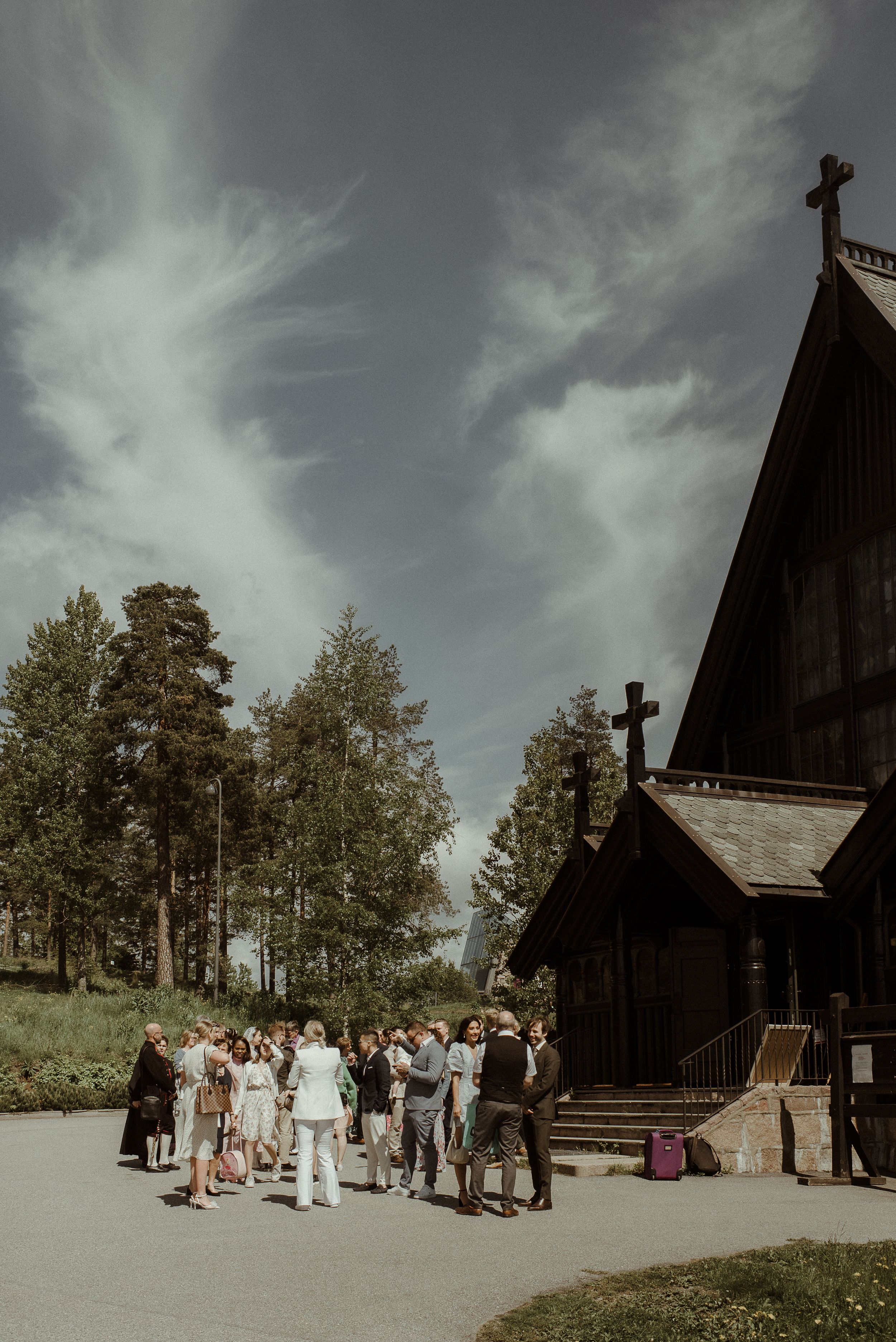 A wooden church with crosses stands against dramatic clouds as guests gather outside.