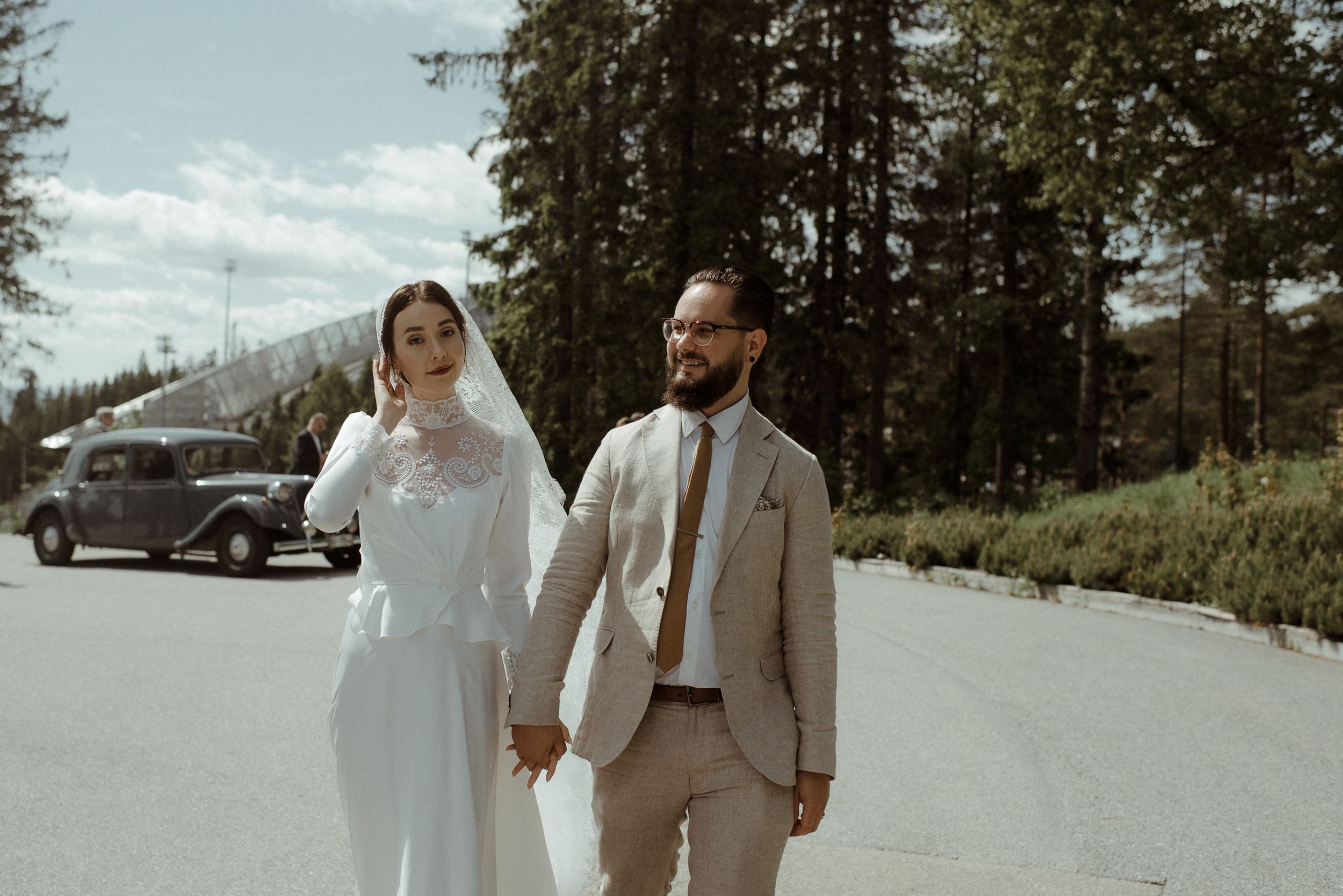 Newlyweds hold hands while walking along a road with vintage car and forest backdrop.