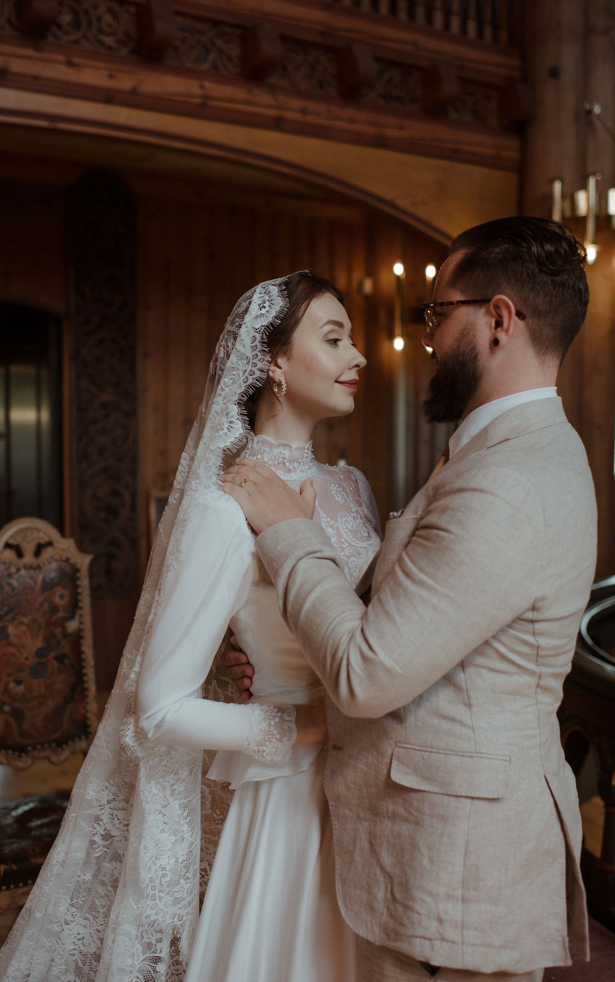 A tender wedding portrait showing the bride in lace and the groom in a light suit against an ornate backdrop.