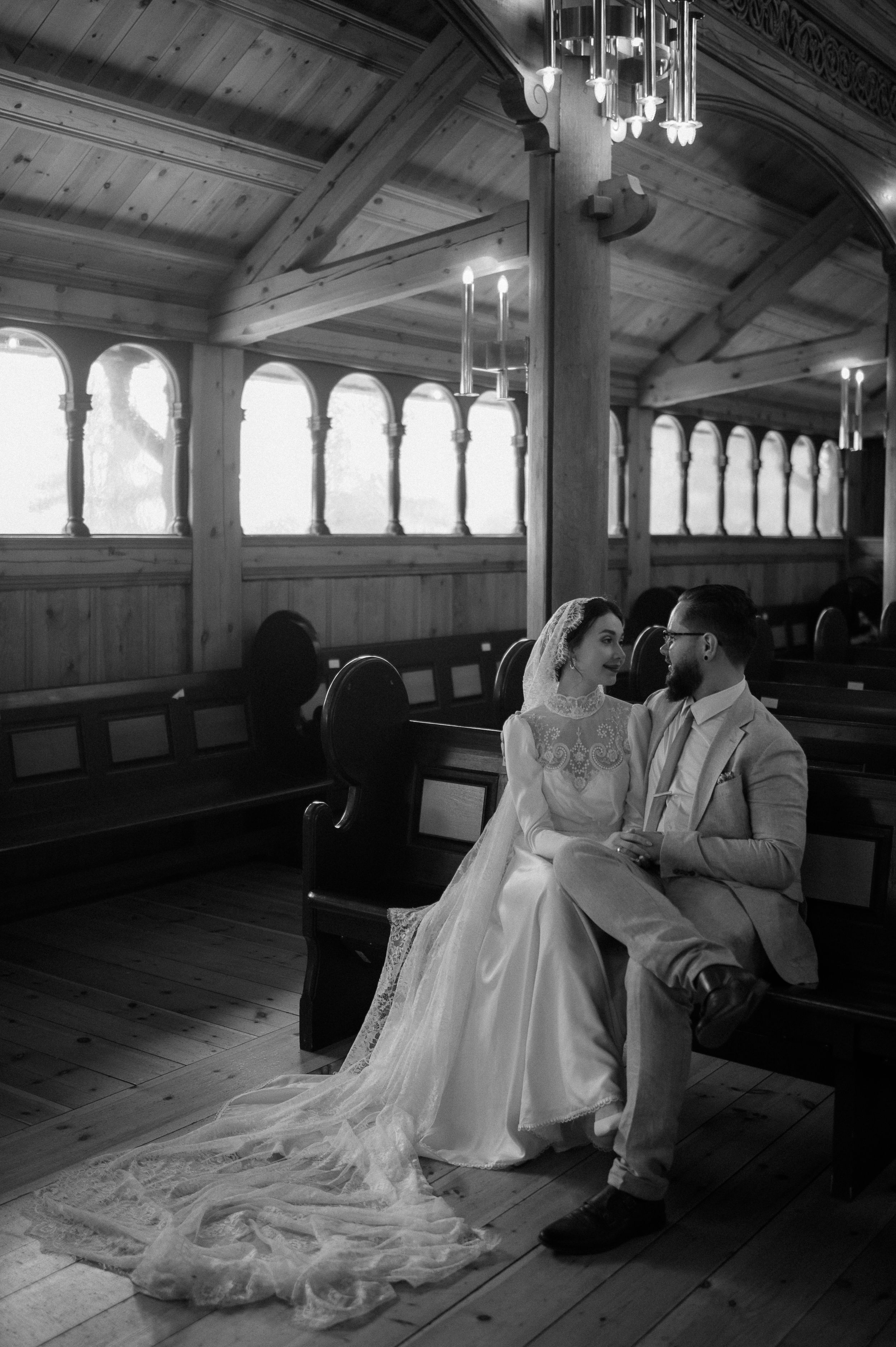 Couple in wedding attire sit in ornate church interior with arched windows and wooden details.