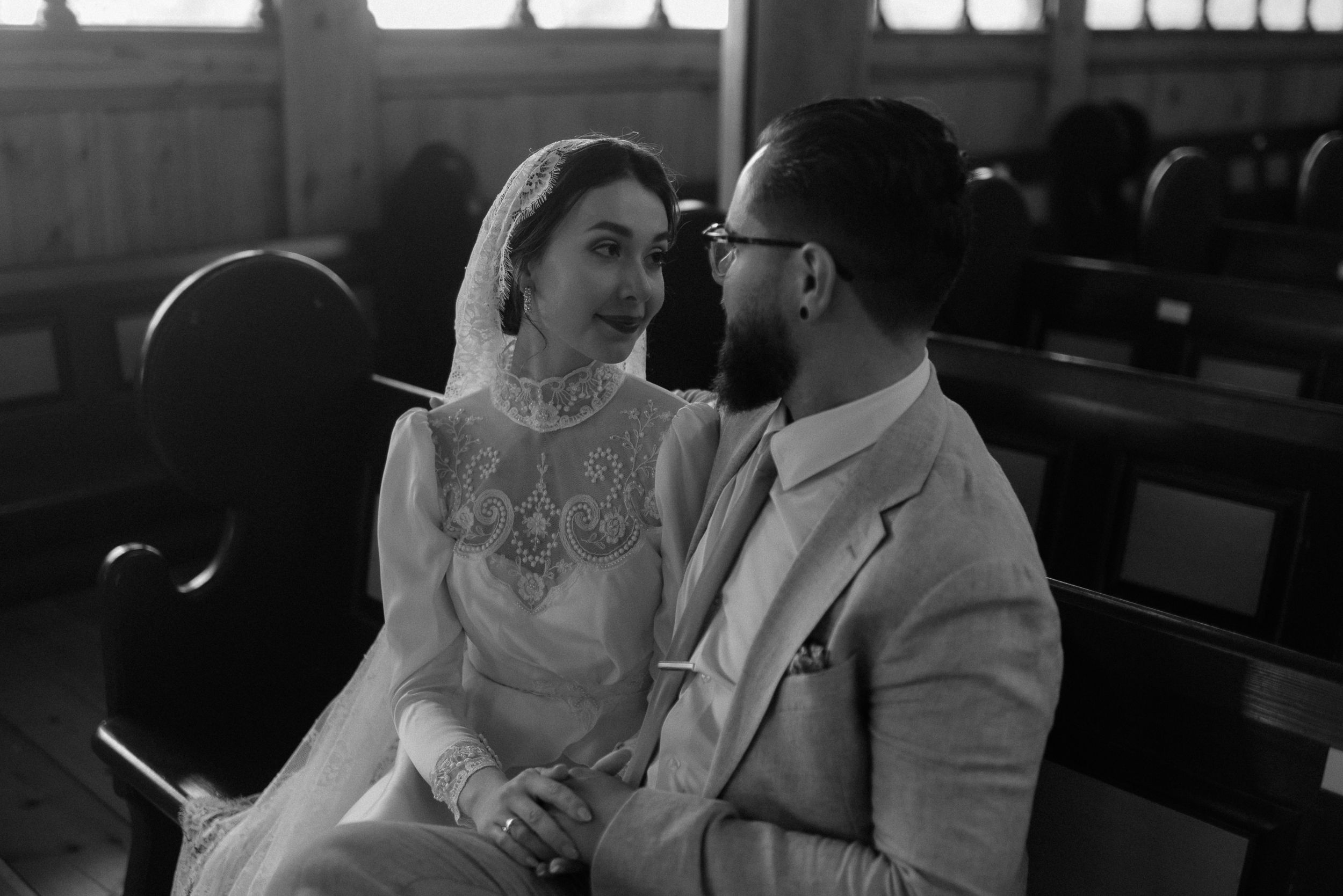 Intimate black and white portrait of couple sitting in church pews on wedding day.