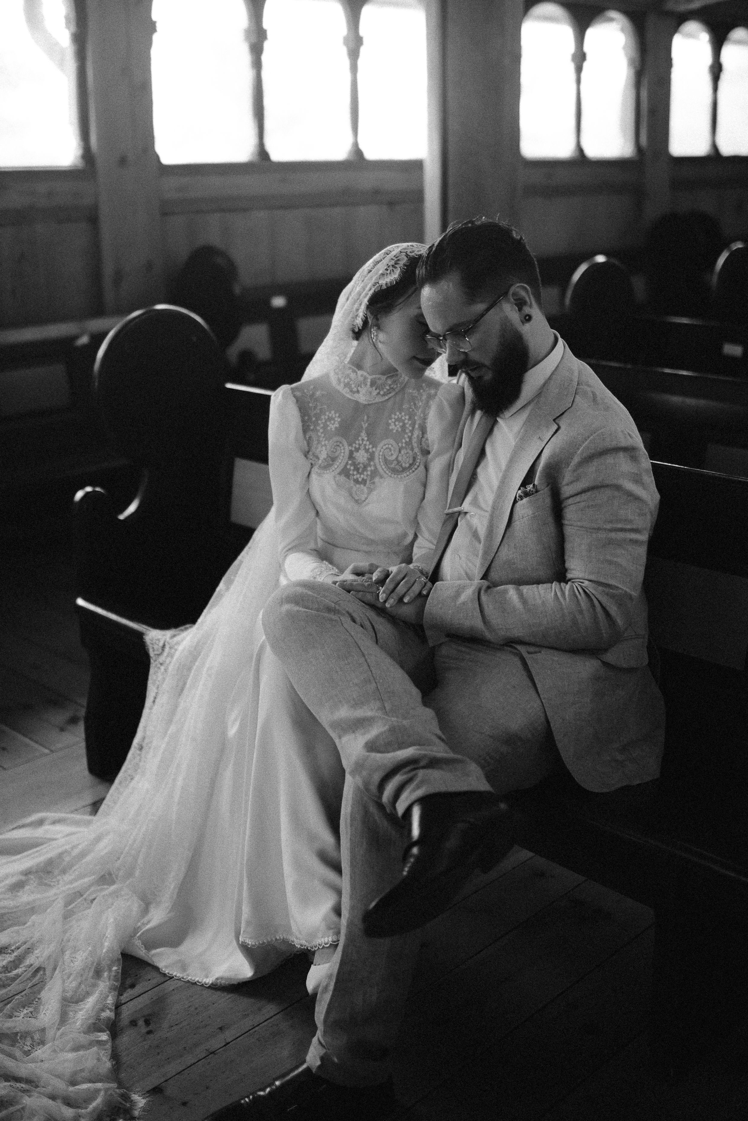Black and white photo of couple sitting intimately in church pew wearing wedding attire.