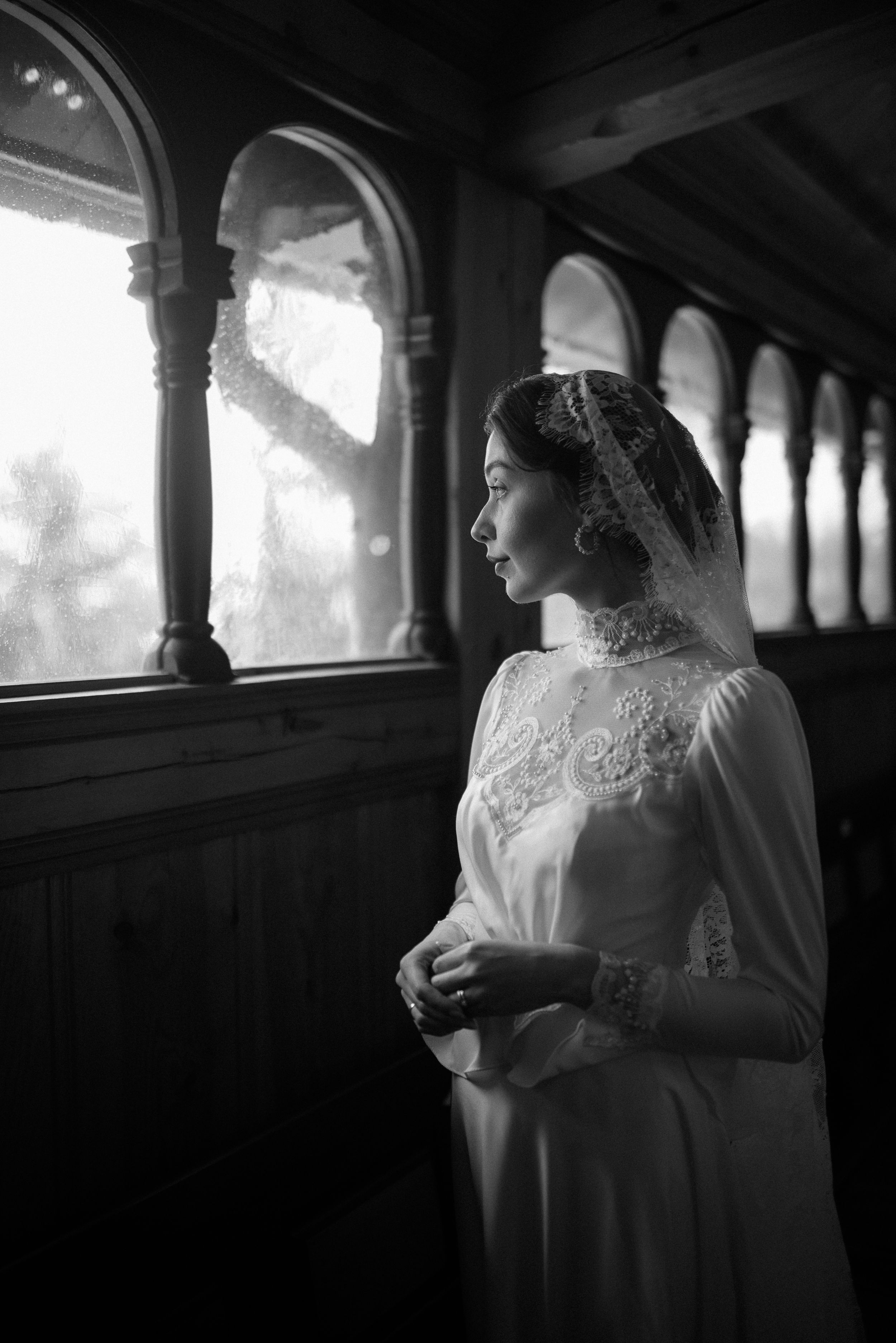 Black and white portrait of bride in lace dress gazing through arched church windows.