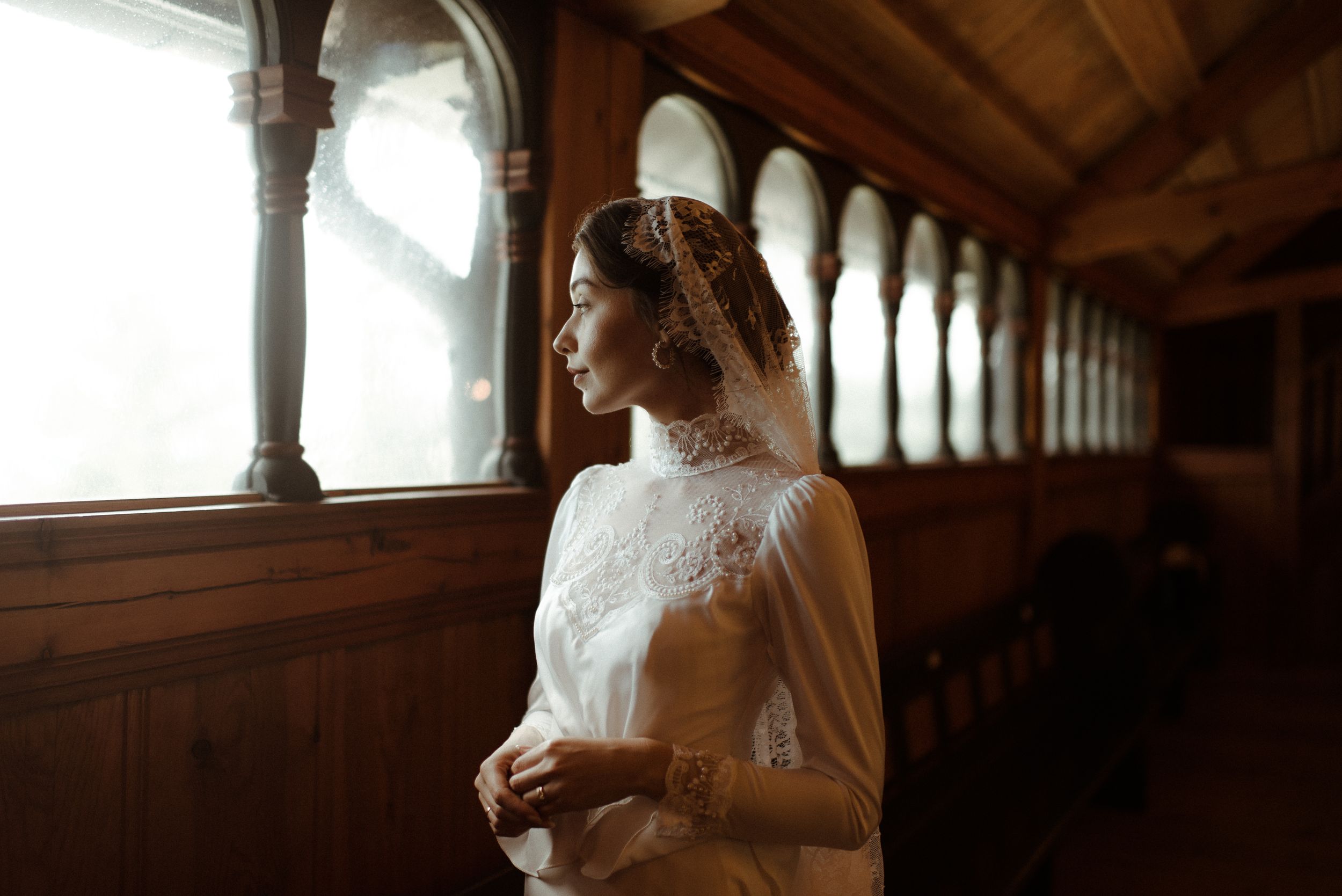 A bride in a white lace gown stands beside ornate wooden archways in warm natural light.