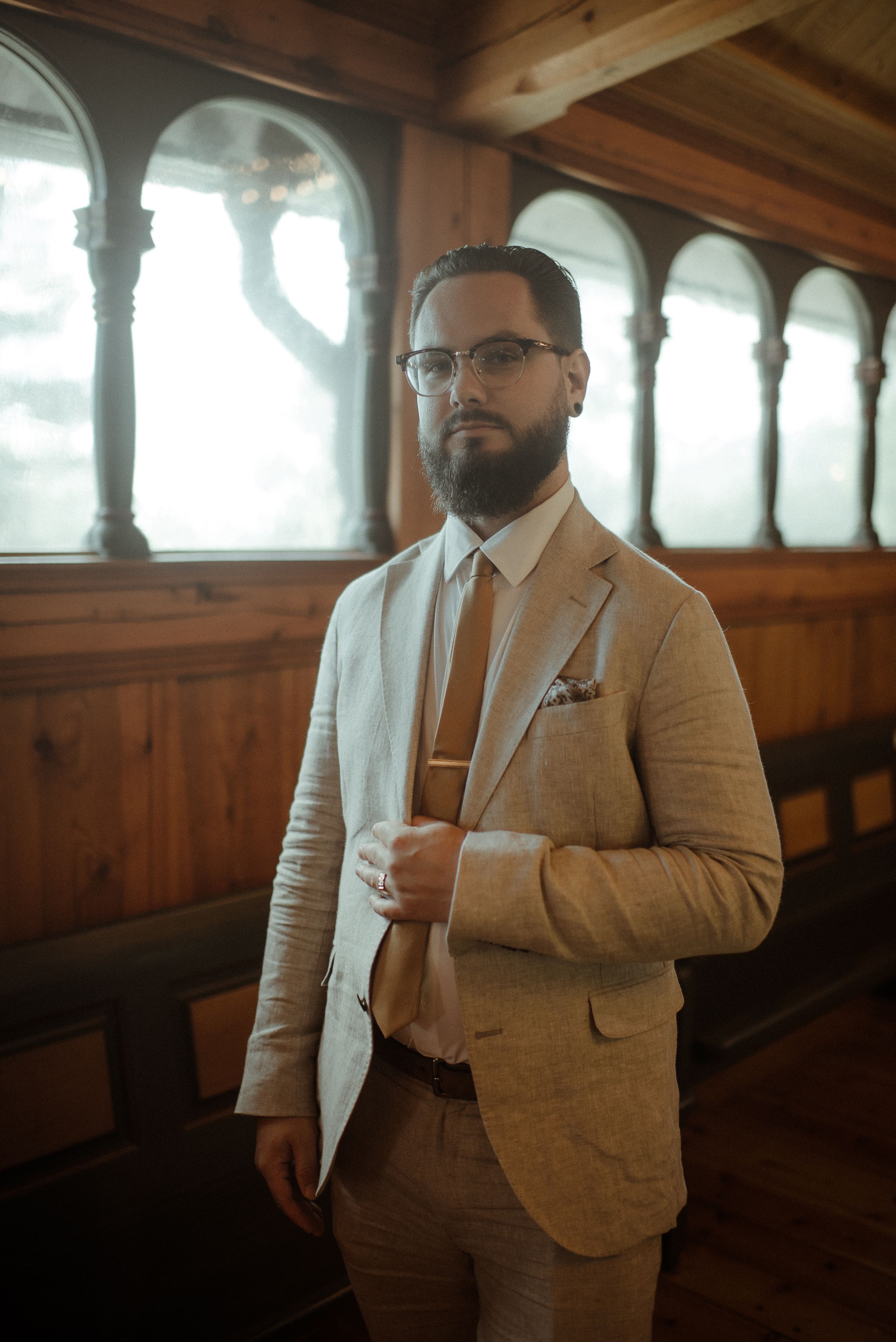 A well-dressed person in a tan suit stands in an elegant wooden archway interior.