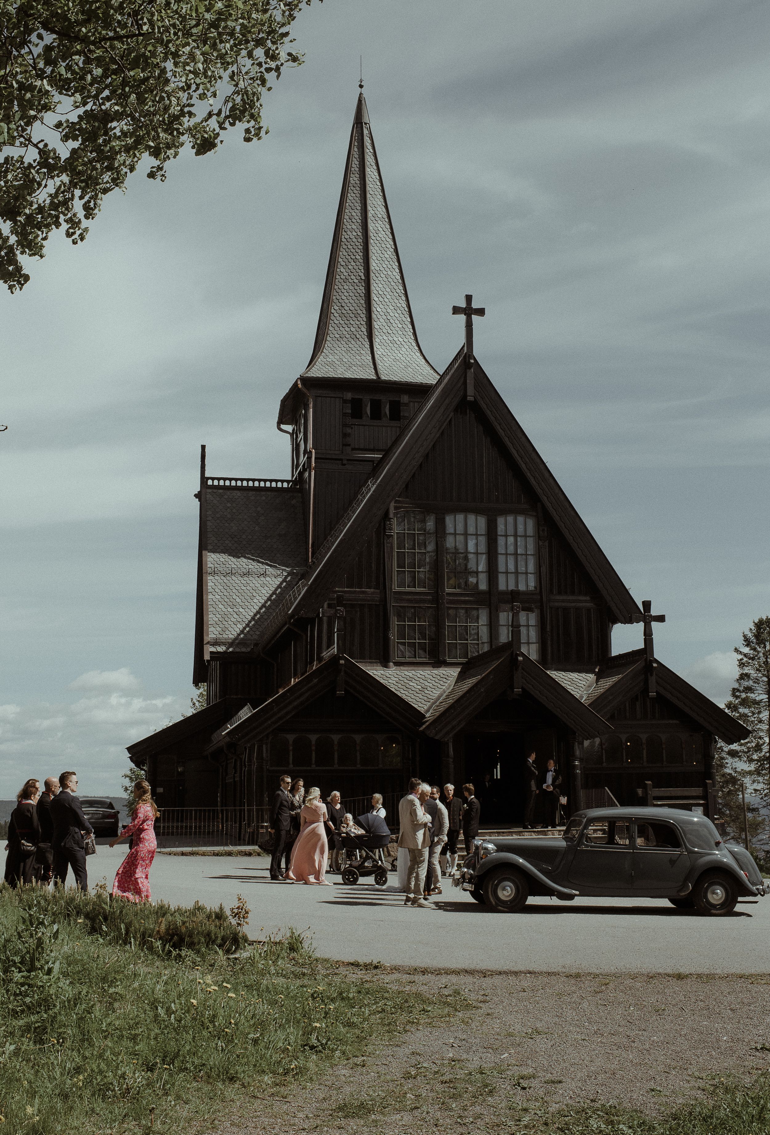 Vintage-style photo of a wooden church with guests gathering outside near classic cars.