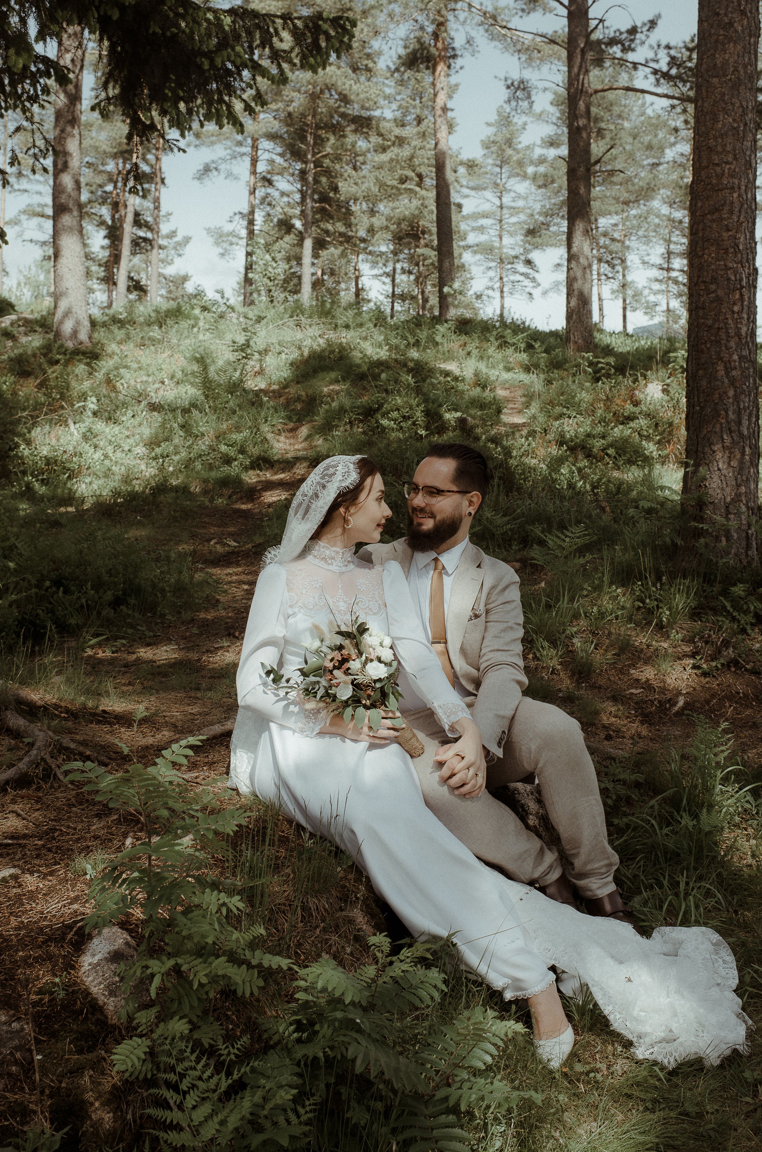Wedding couple sits intimately on forest ground surrounded by tall pine trees.