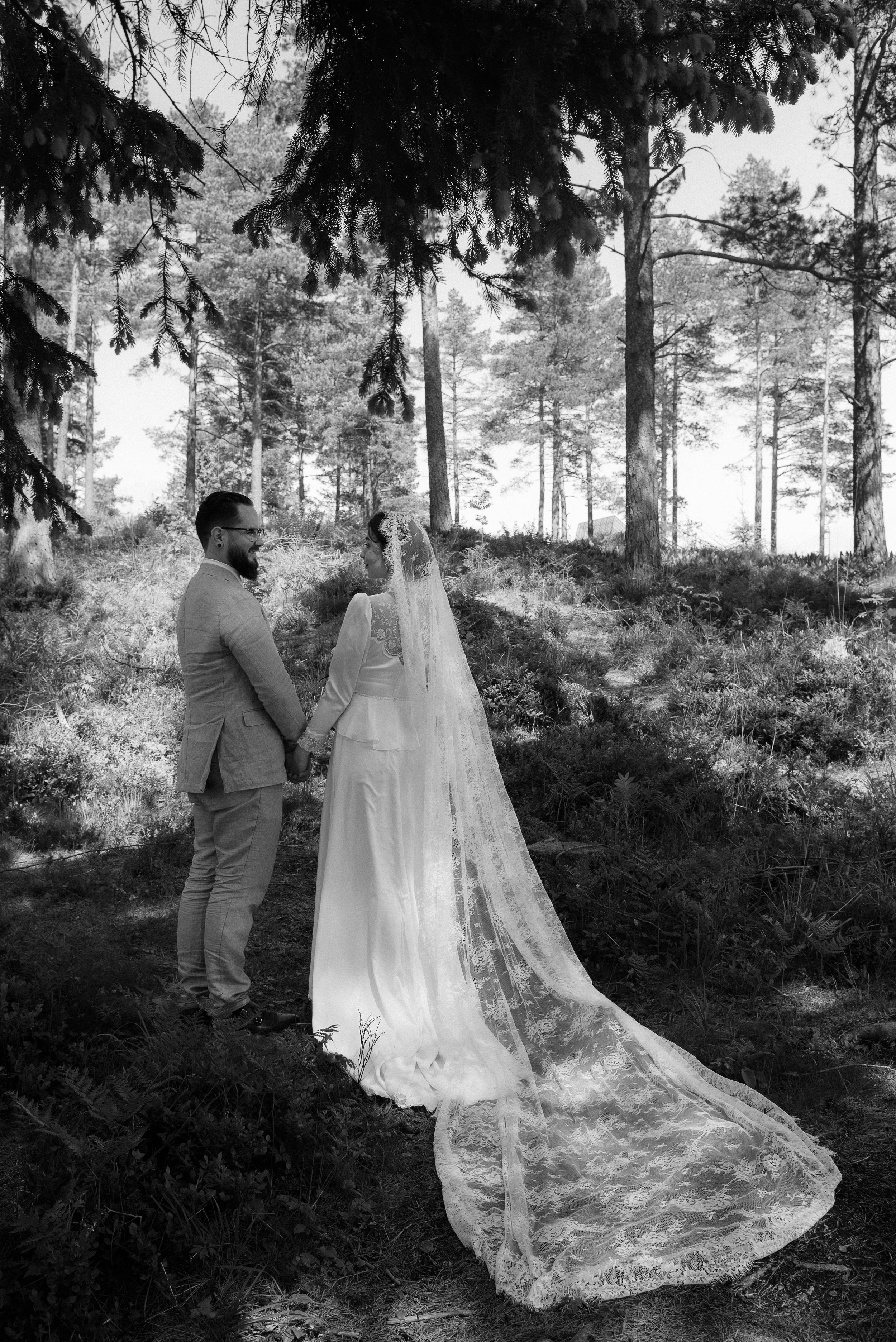 A wedding couple poses in a forest setting with bride's dramatic lace veil trailing behind.