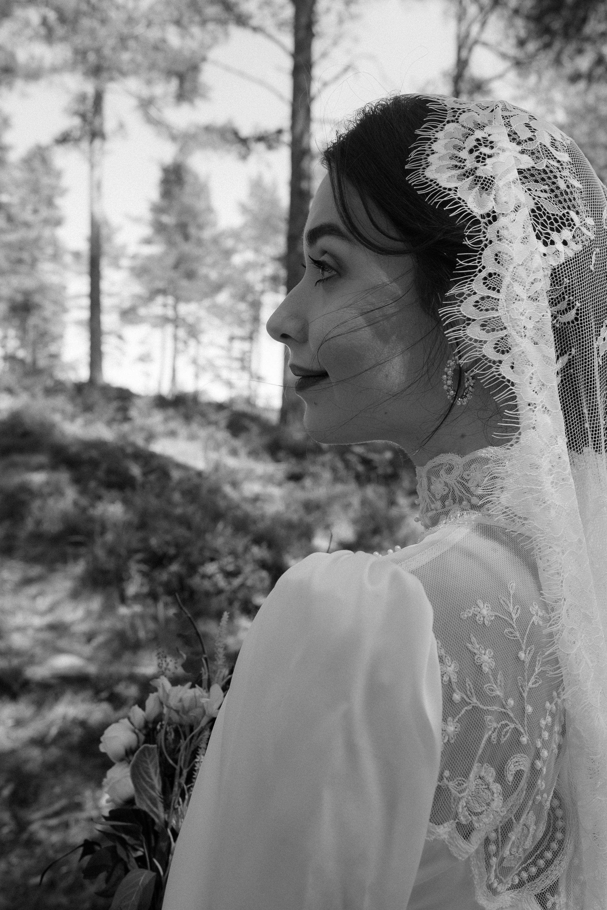 Side profile of bride wearing delicate lace veil against forest backdrop.