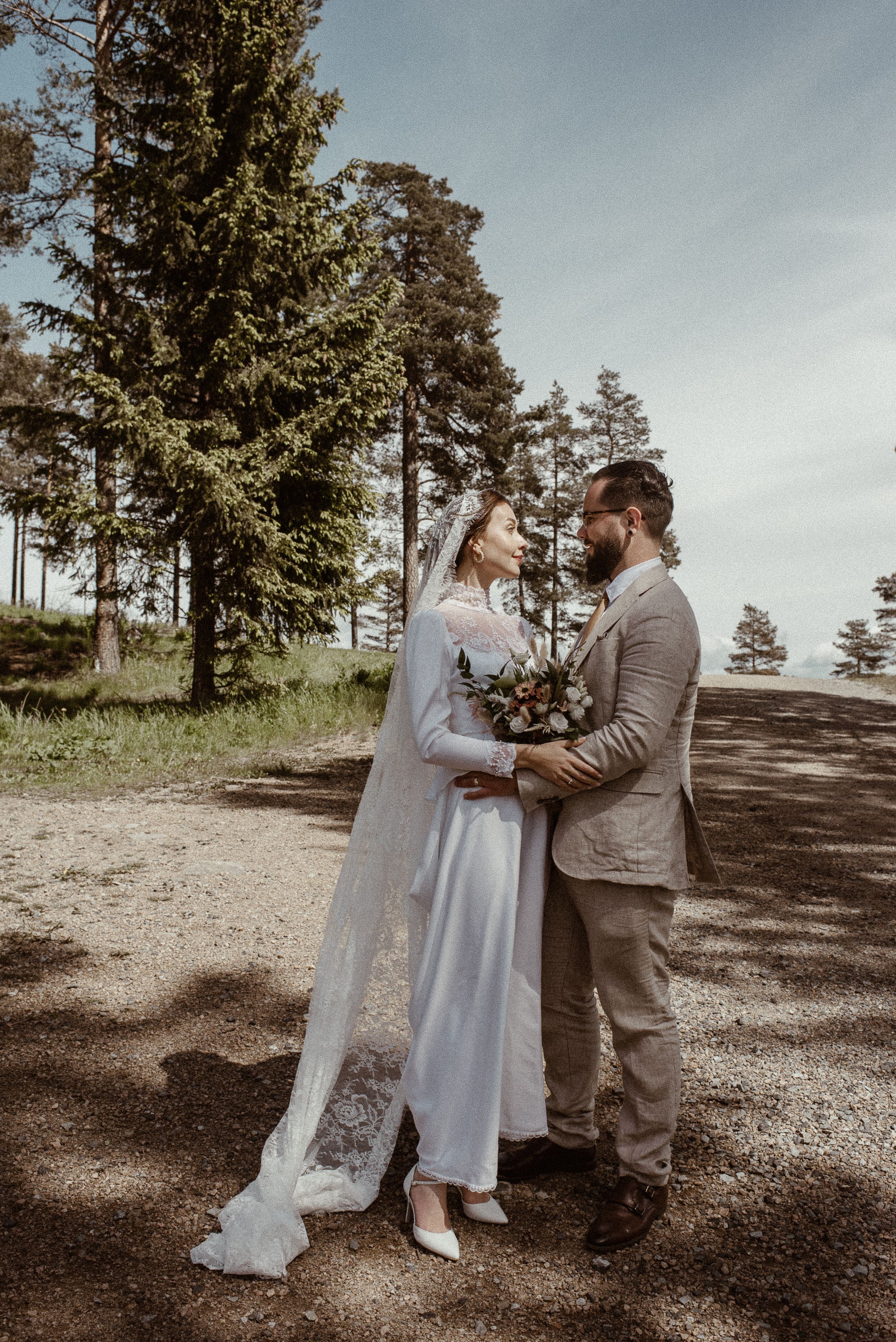 Wedding couple embraces on forest path with tall pine trees in background.