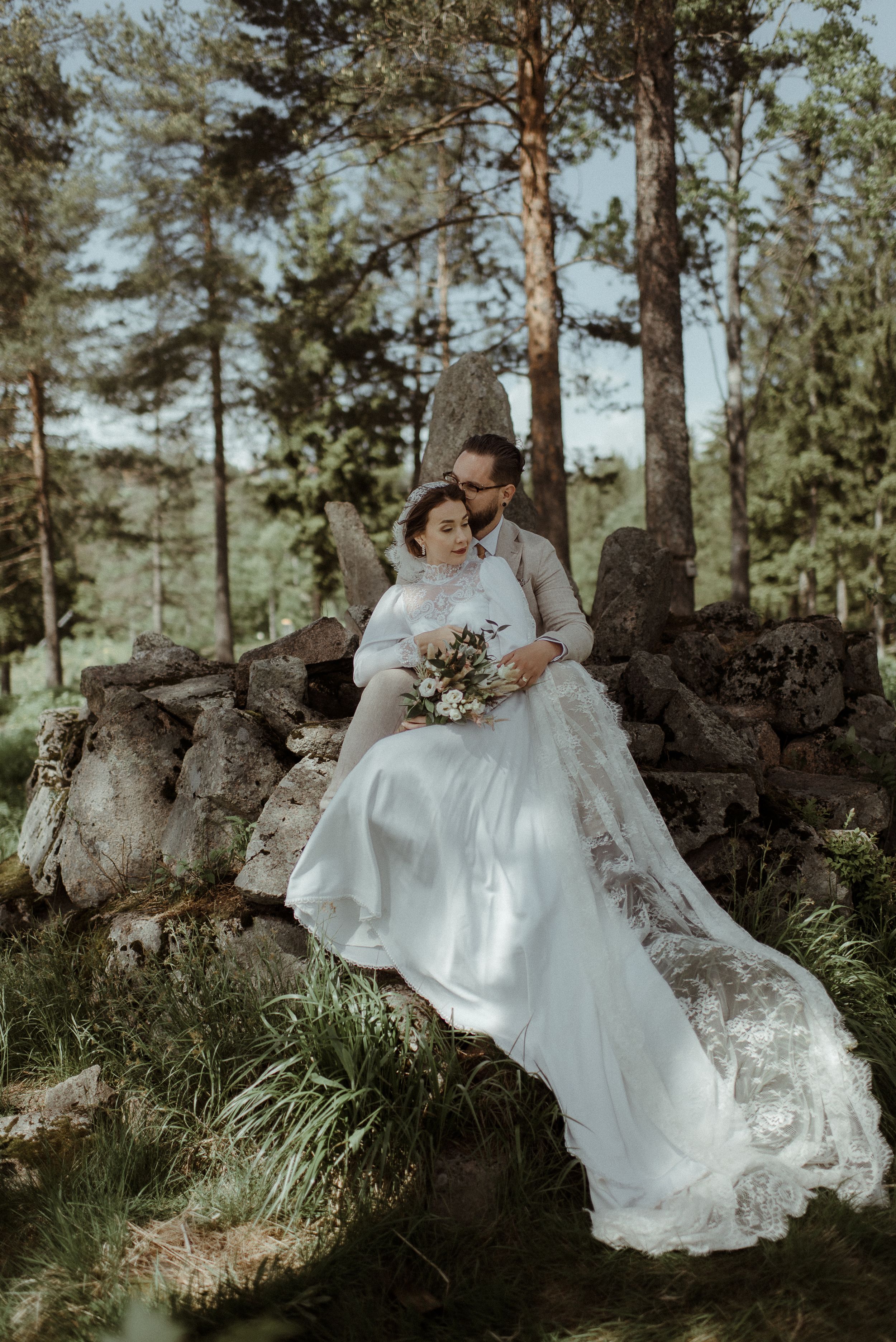 Romantic wedding portrait of couple sitting on rocky outcrop in sunlit forest setting.