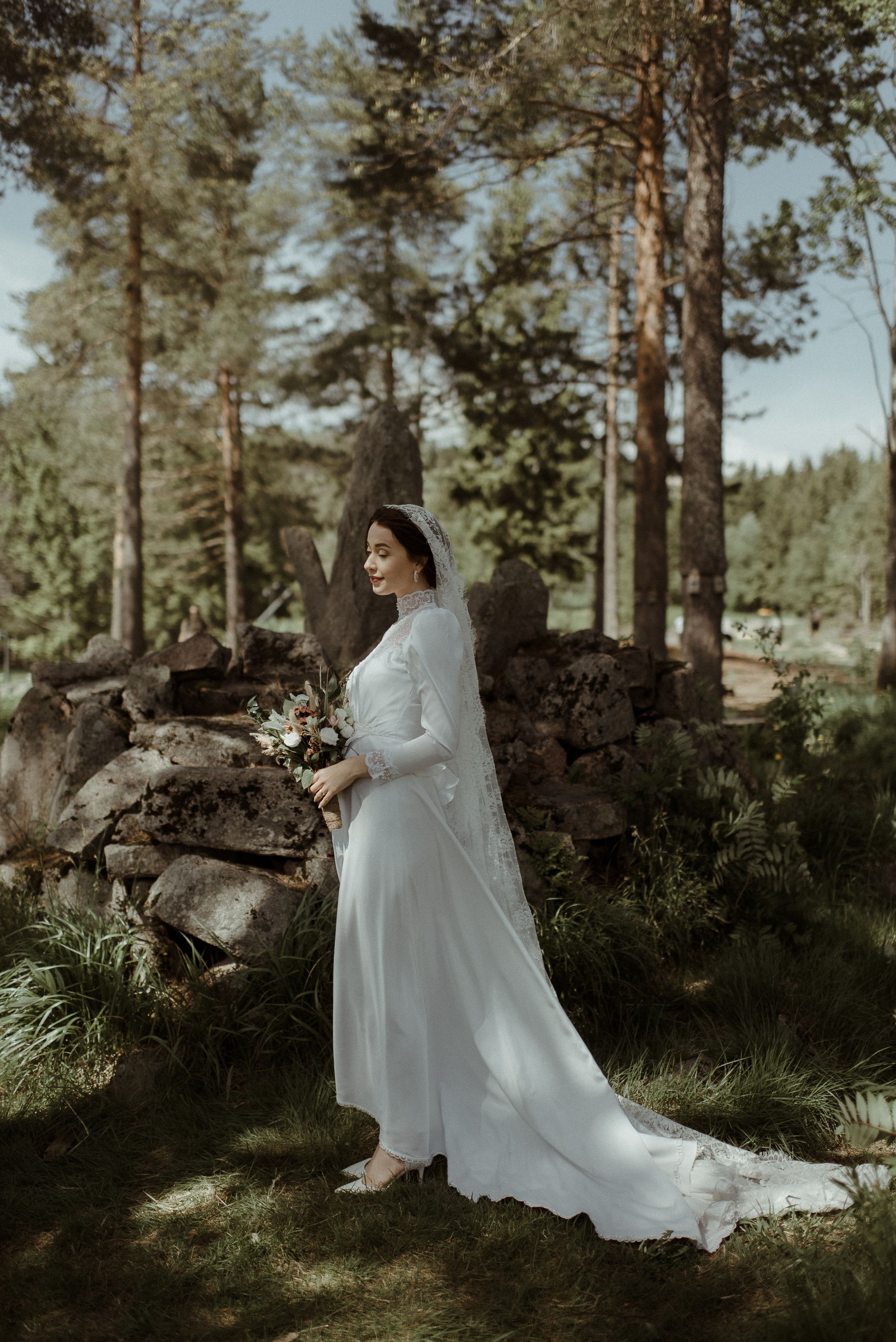 A bride in a long-sleeve white gown poses gracefully in a forest setting with tall pine trees.