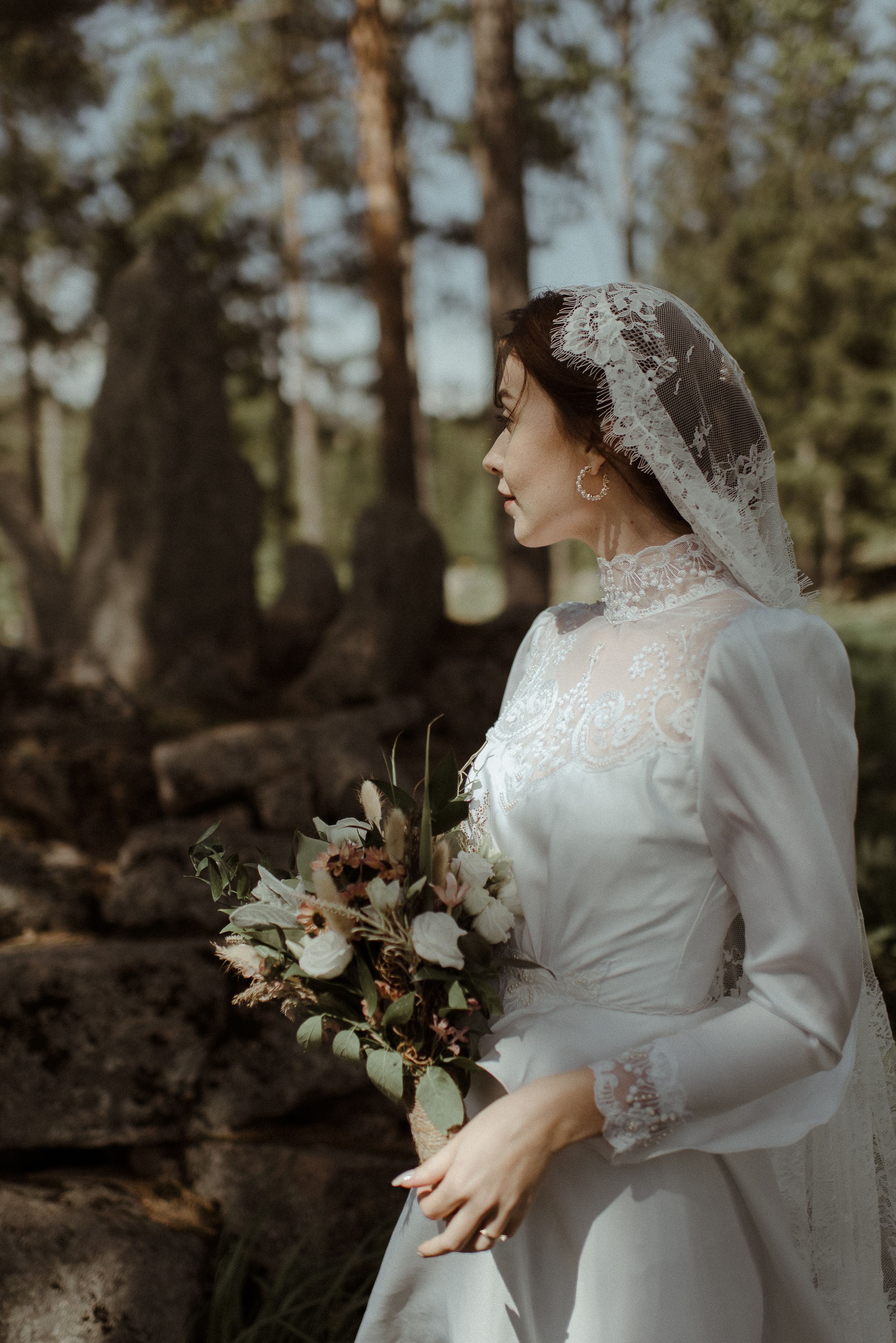 Elegant bride in high-neck lace gown holds bouquet of mixed flowers in forest setting.