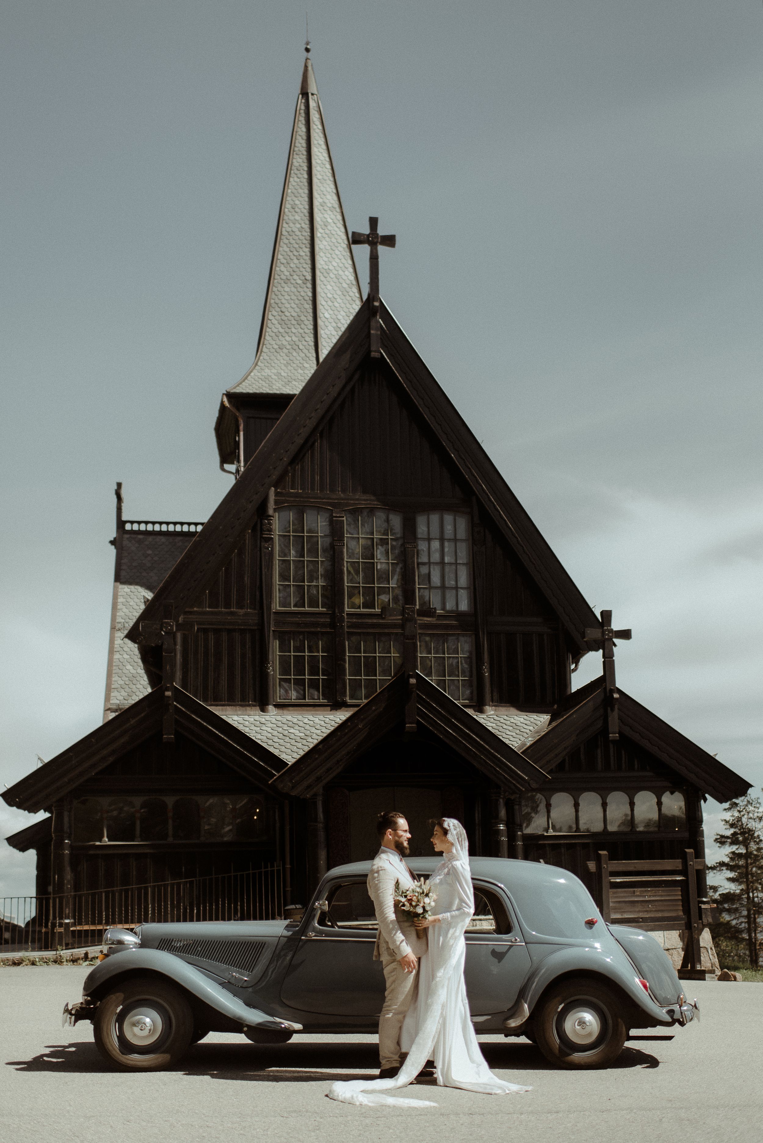 Classic vintage car parked in front of wooden church with dramatic spire against moody sky.