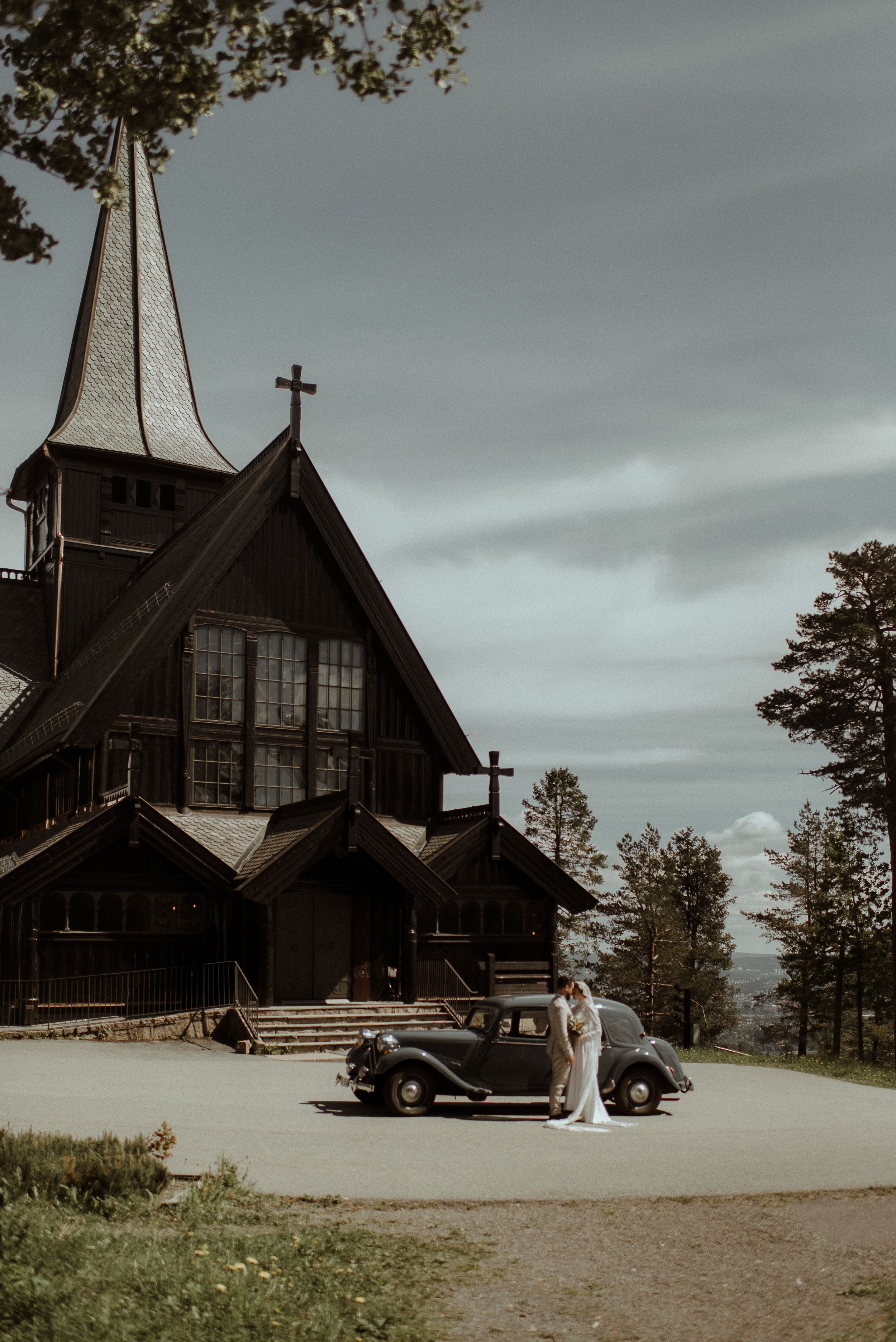 Wedding scene with classic car and wooden church architecture against cloudy sky.
