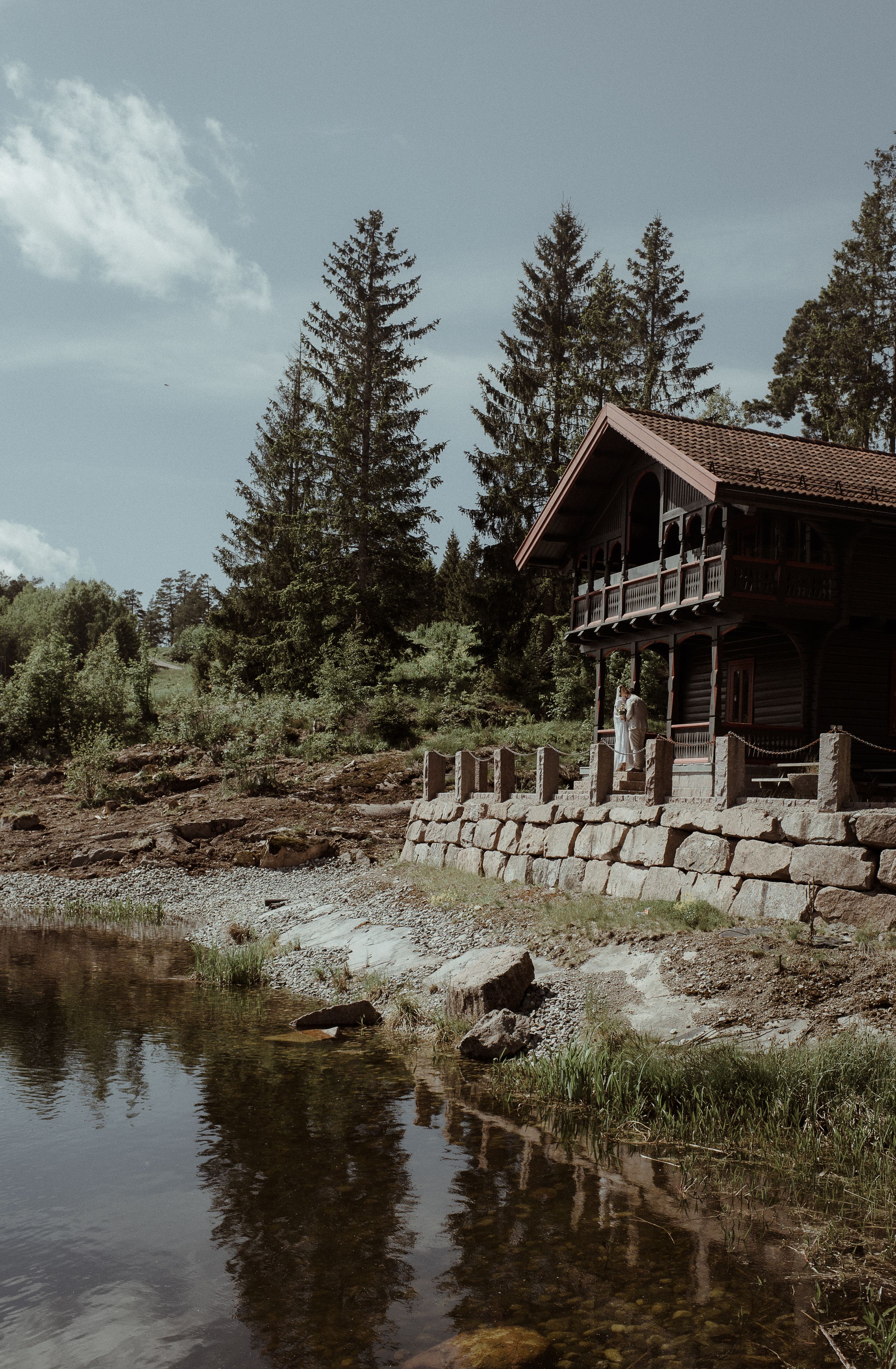 A rustic wooden cabin with a balcony sits beside a tranquil lake surrounded by evergreen trees.