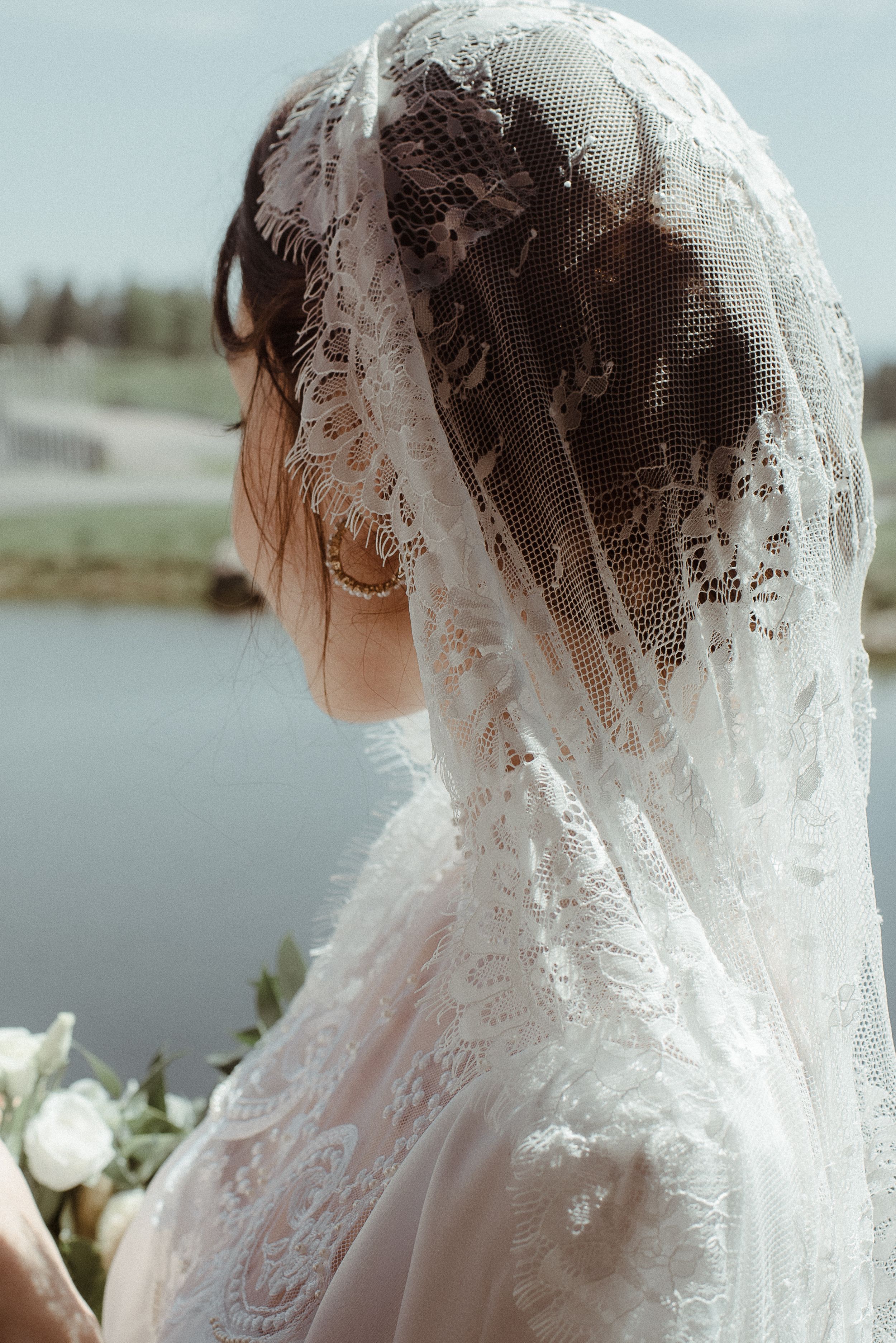 A close-up side view of an elegant lace wedding veil against a waterfront backdrop.
