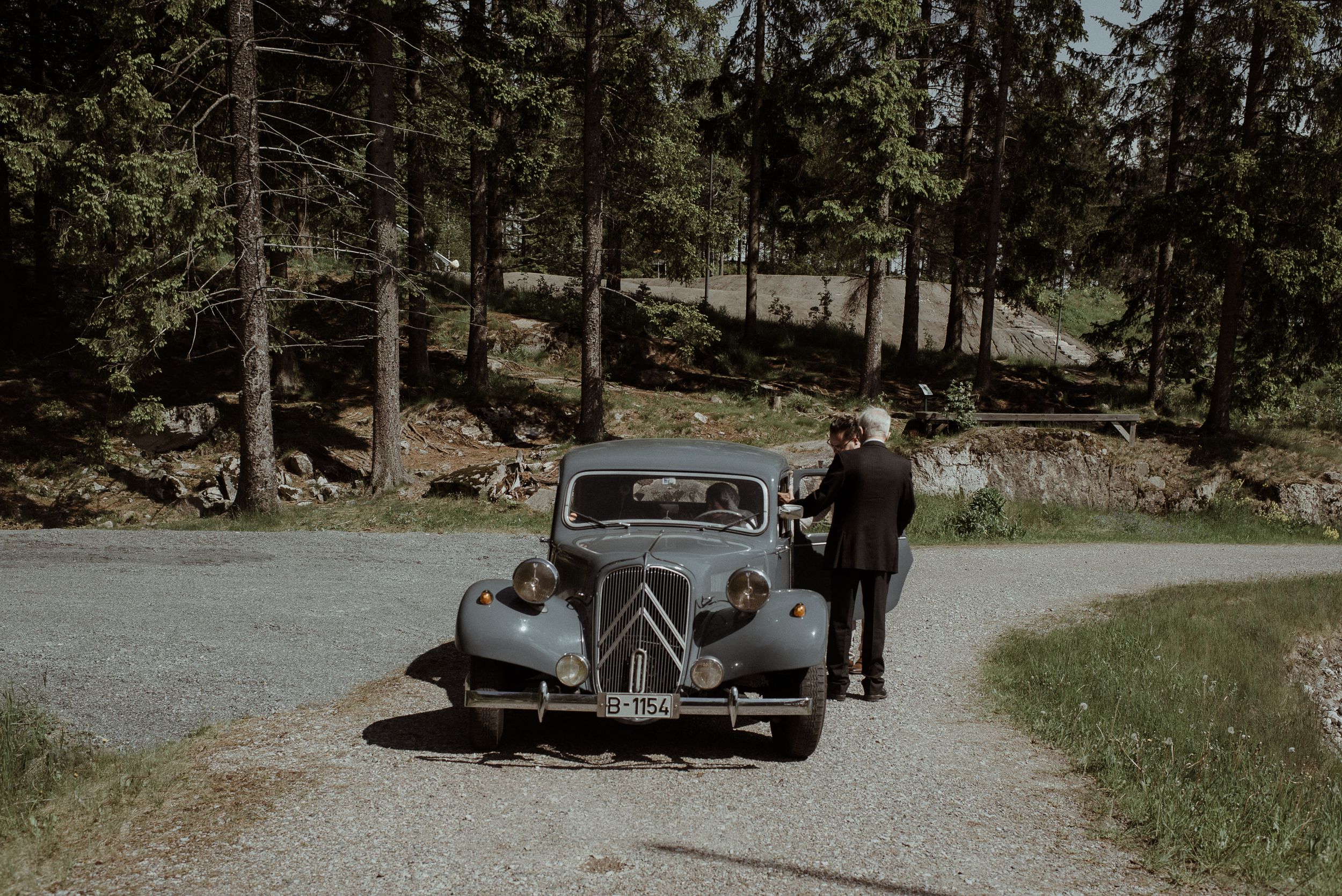 A classic vintage car parked on a forest path with wedding attendees.