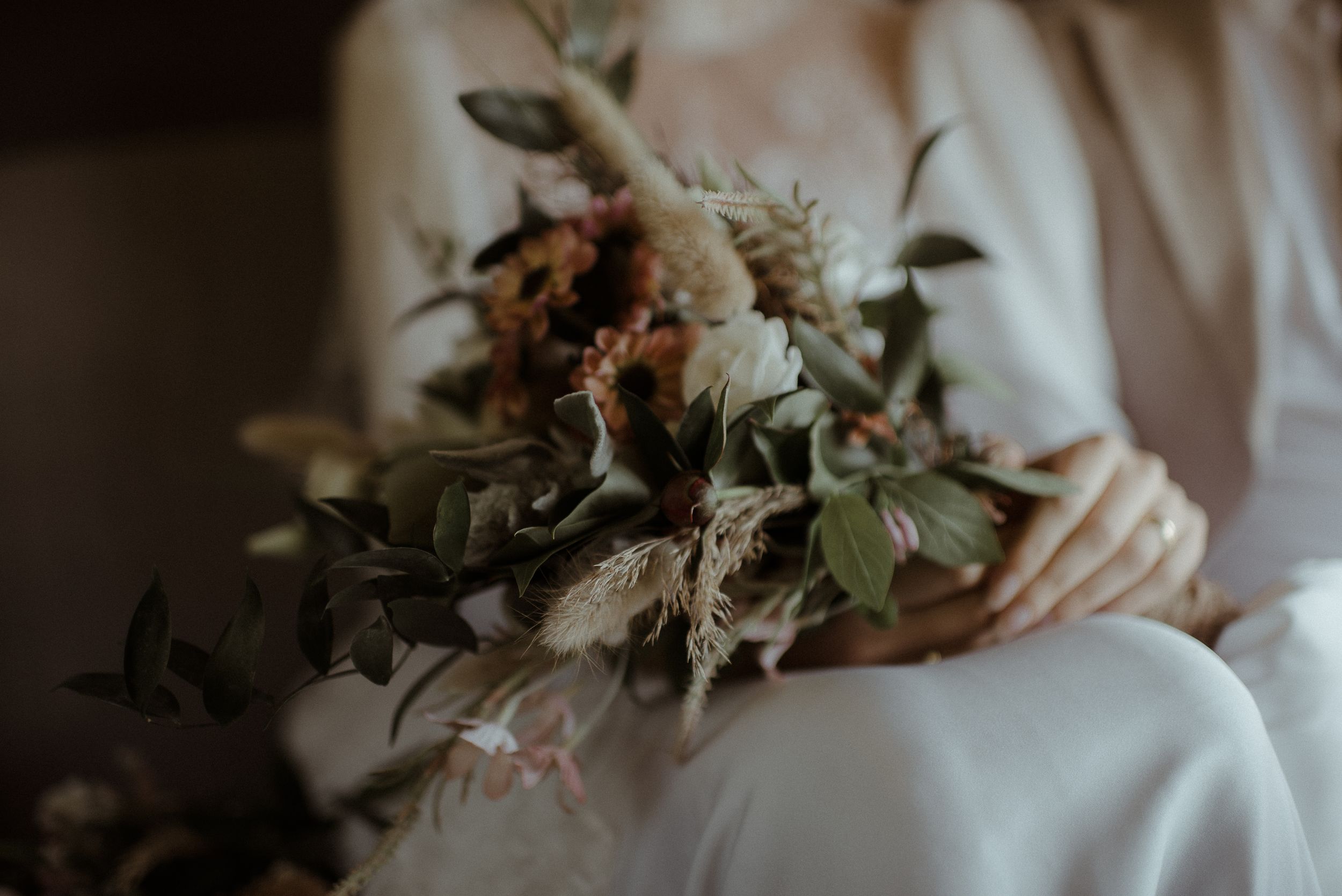 A delicate wedding bouquet with dried flowers and greenery held against a white dress.