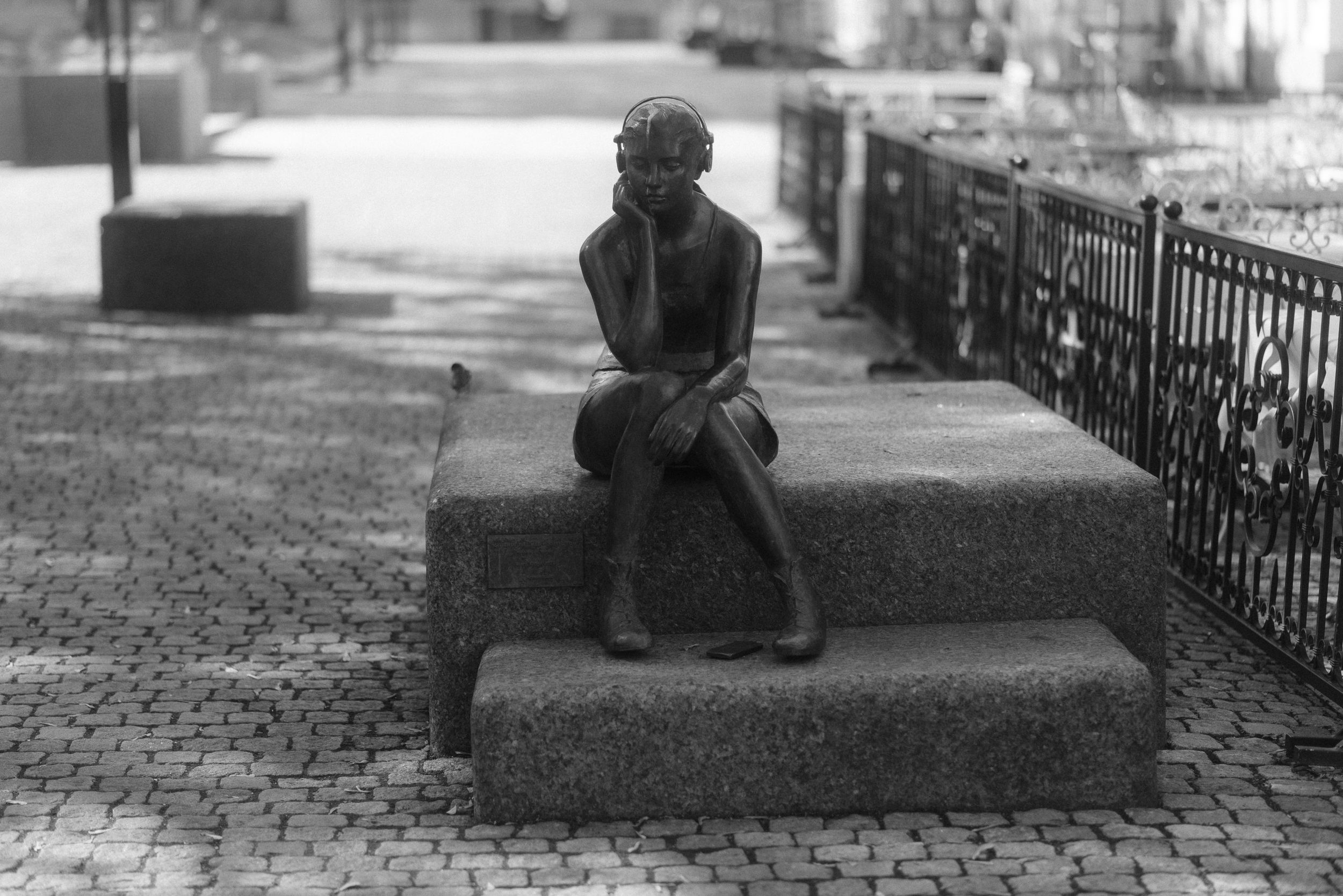 A bronze statue sits thoughtfully on stone steps beside an iron fence.