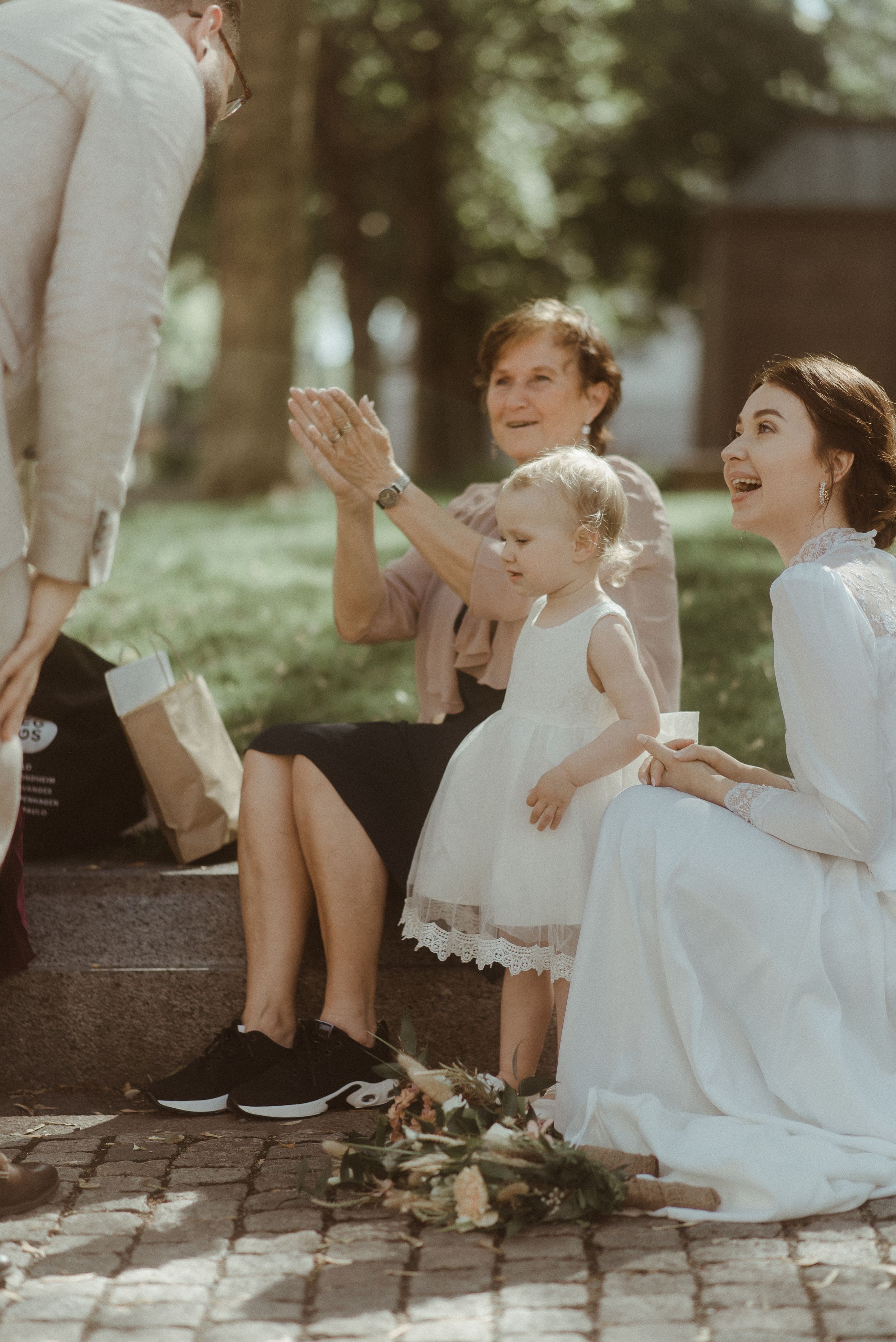 People in wedding attire sit on stone steps during an outdoor celebration.