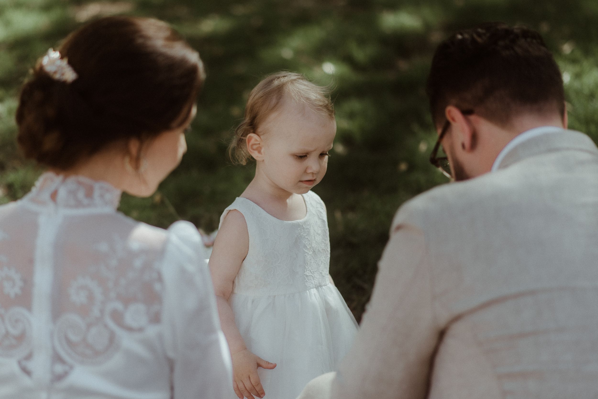 A small child in white dress stands between adults during an outdoor gathering.