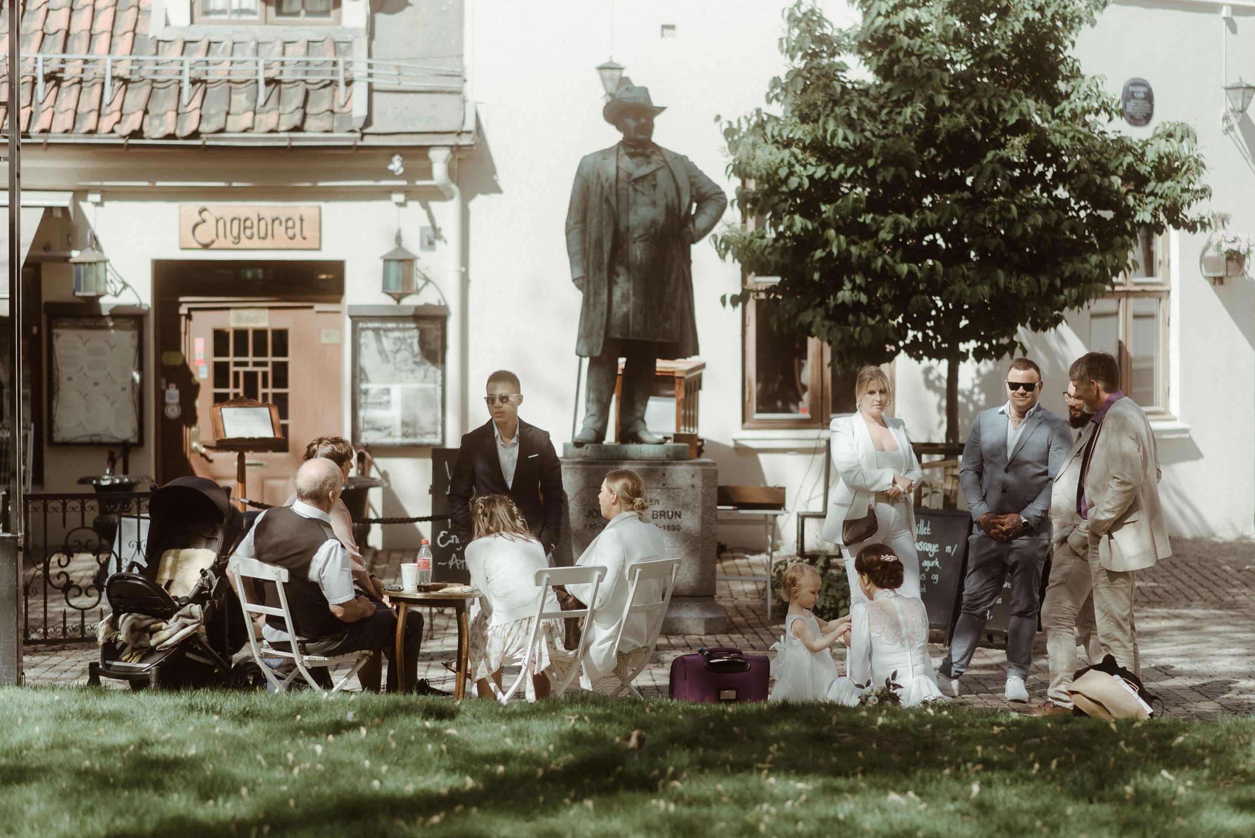 People gathered around a statue in a garden setting with historic architecture in the background.