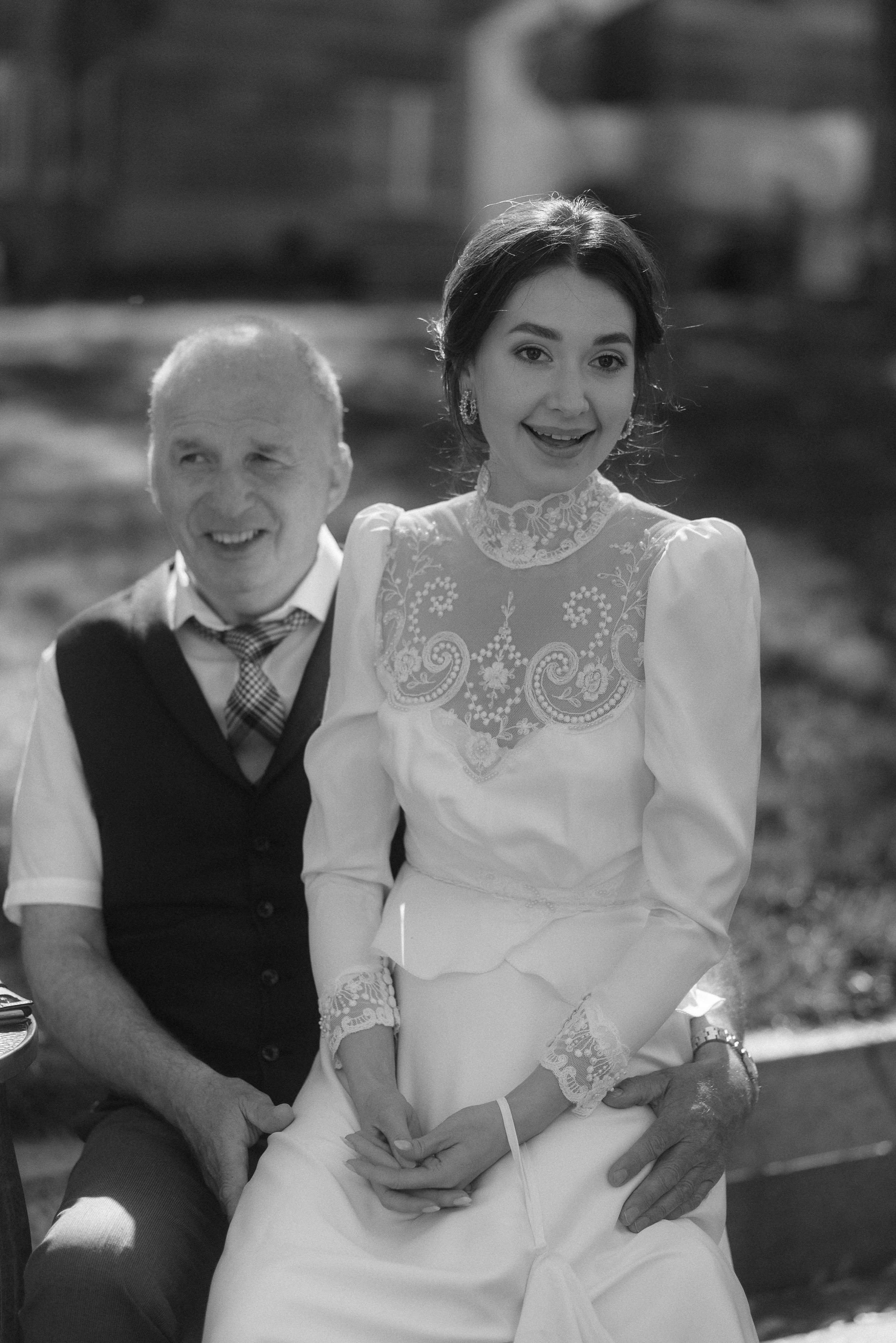 A couple sits together outdoors, with the woman wearing an elegant white lace wedding dress.