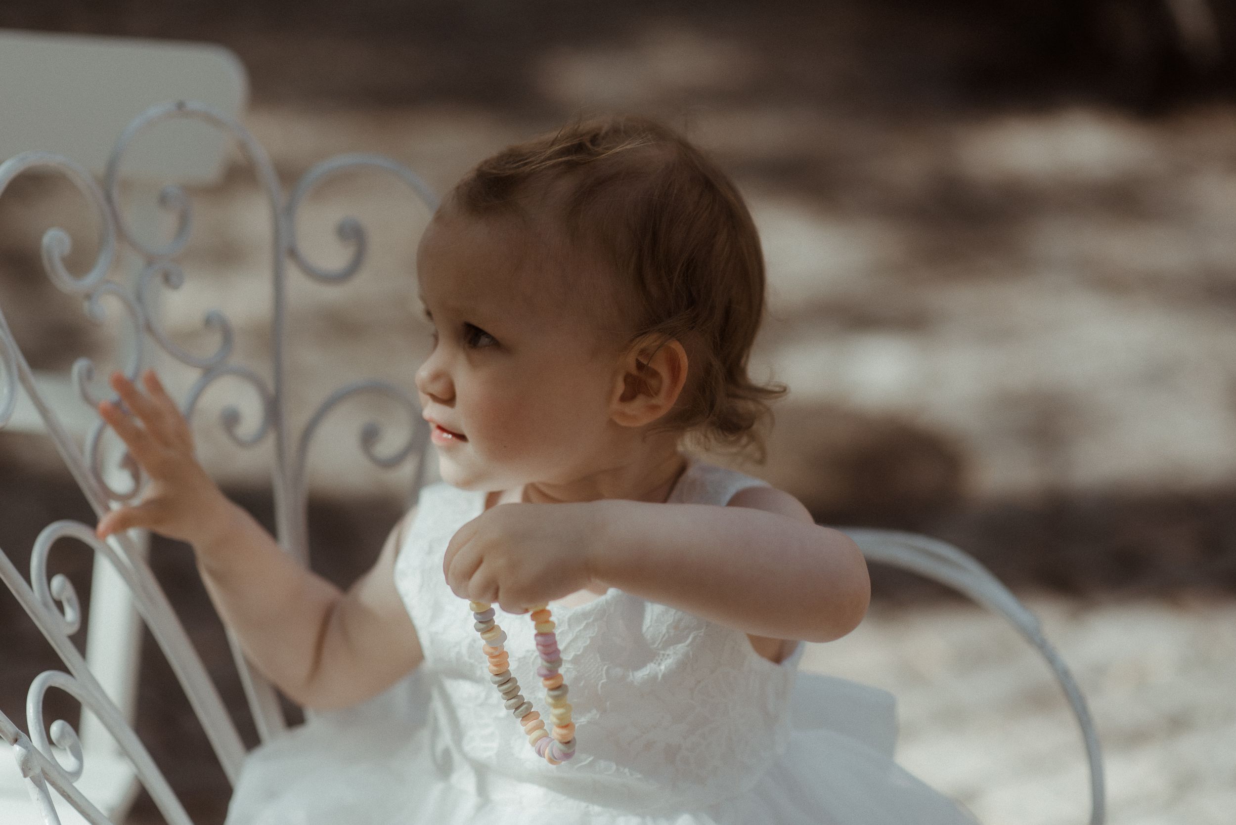 A baby in white dress sits in an ornate white metal chair during an outdoor event.