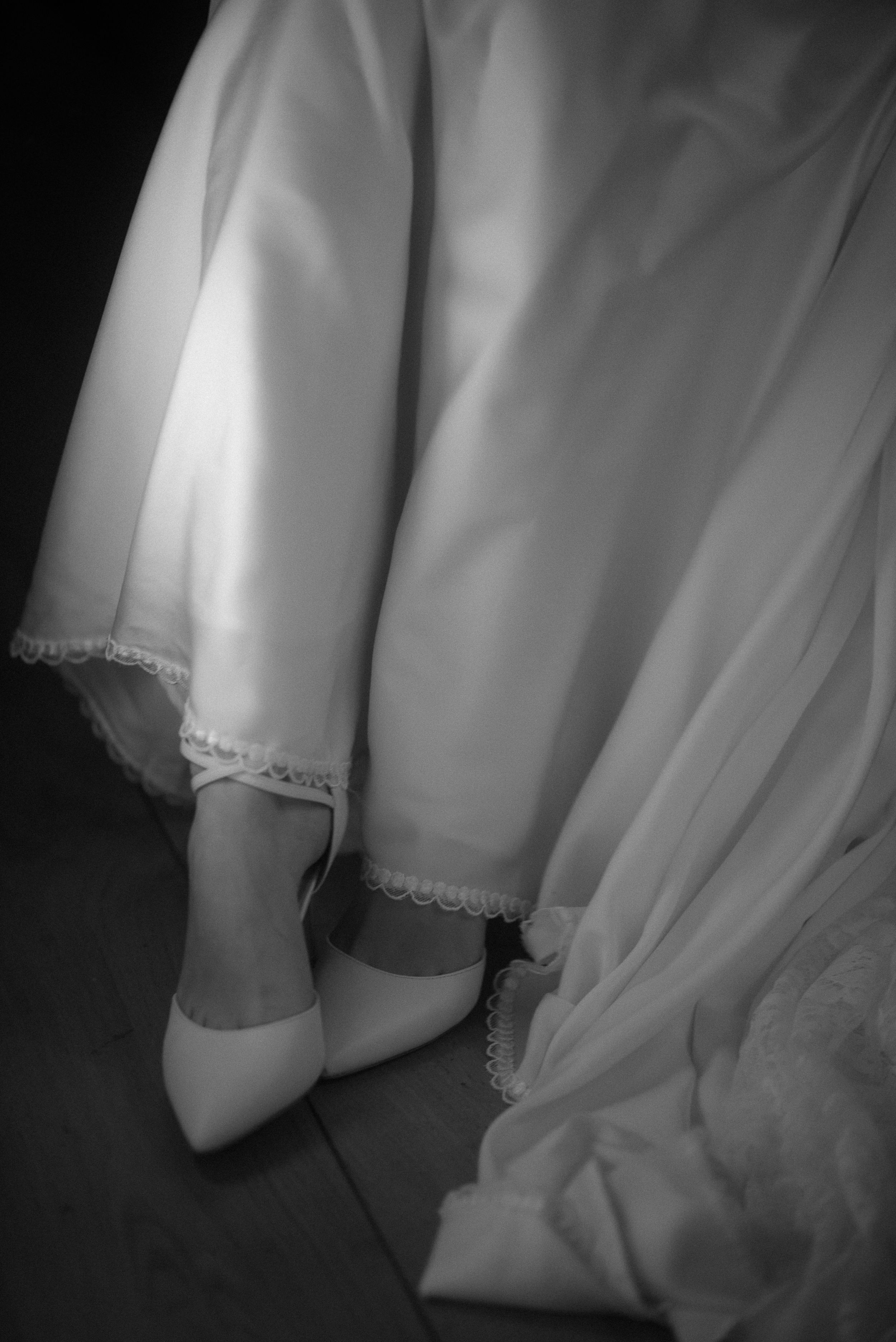 Close-up of white bridal shoes and lace dress hem in black and white photography.