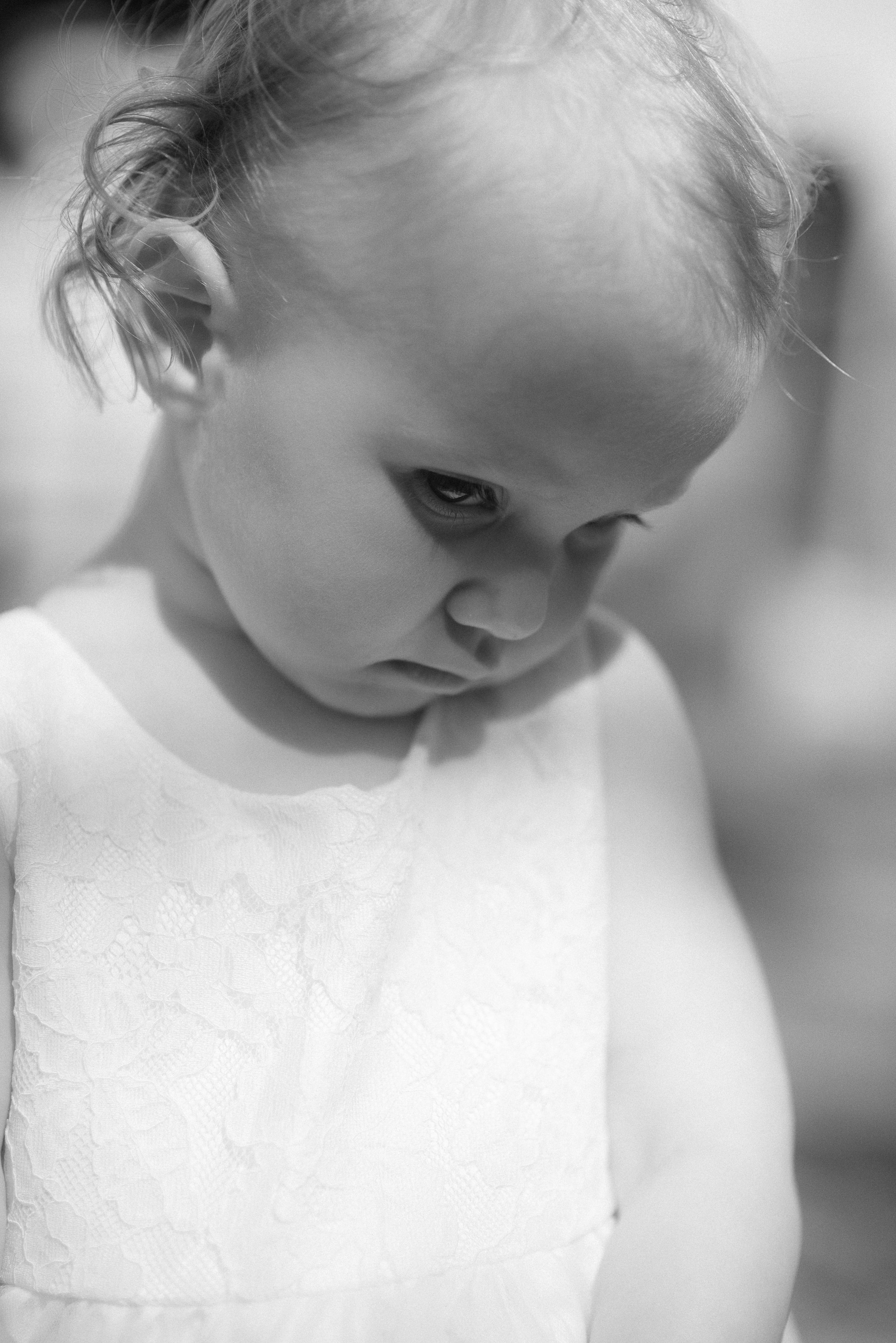 A close-up black and white portrait of a young child in white clothing with a contemplative expression.