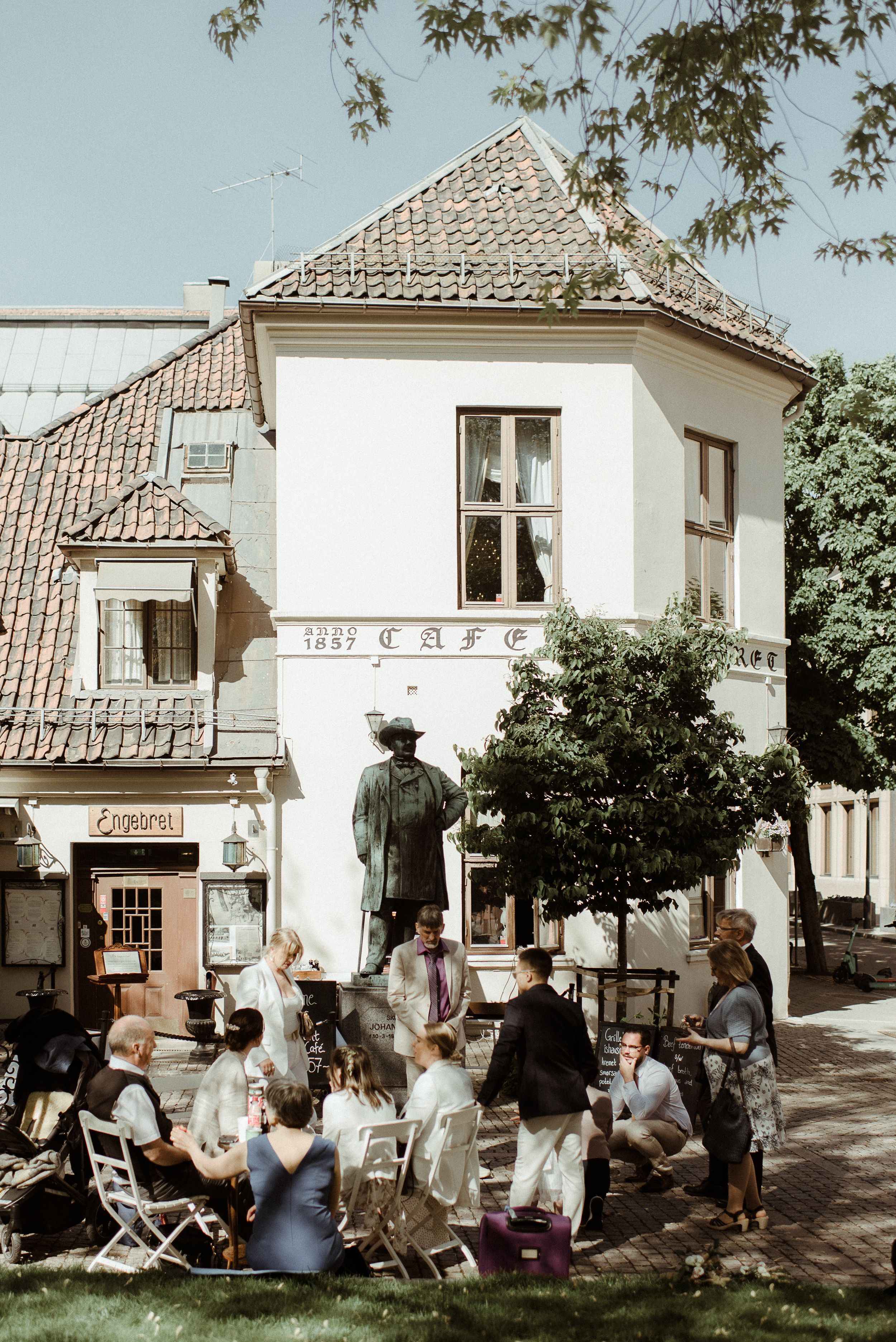 A group of people relaxing outside a quaint white cafe building with a tiled roof on a sunny day.