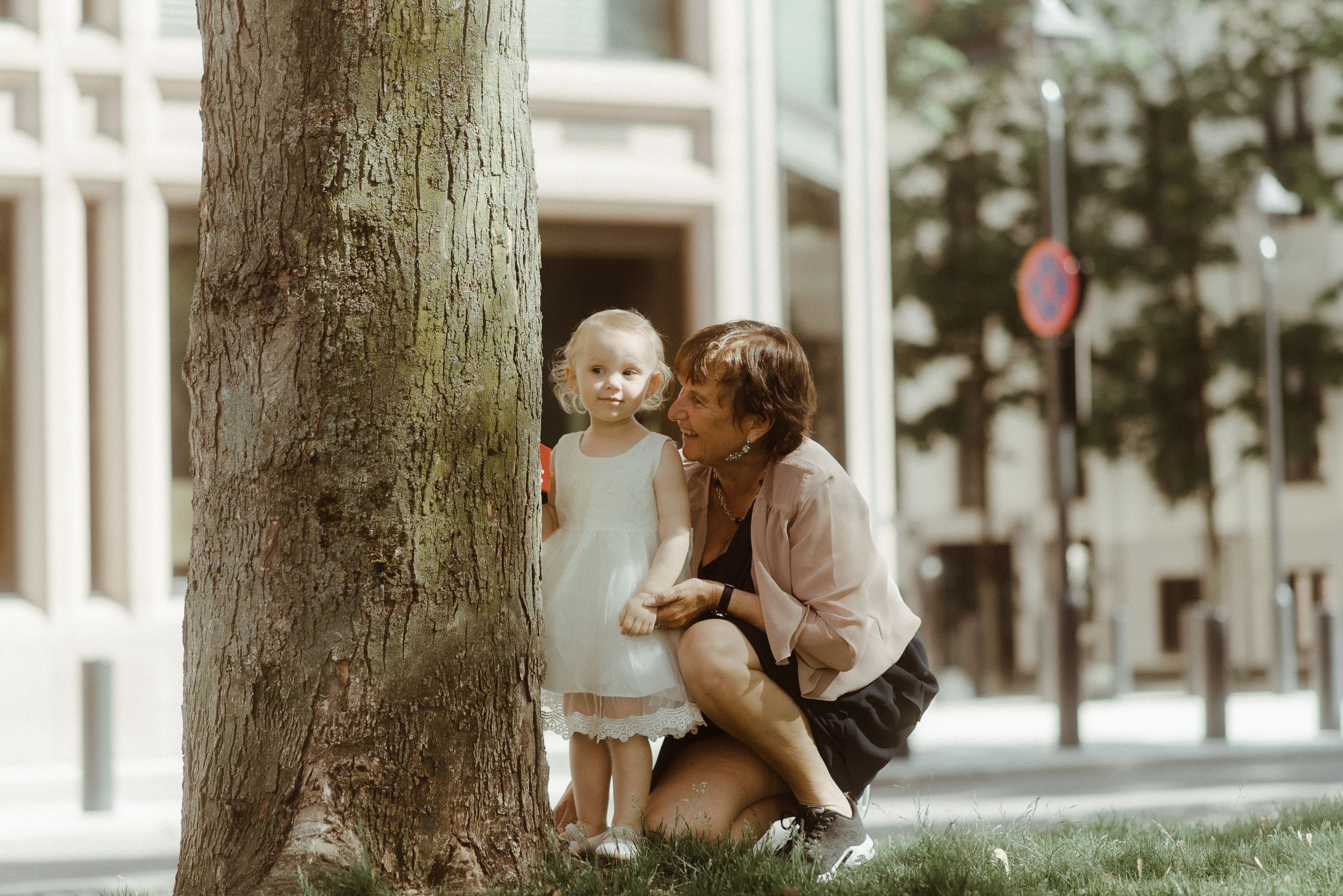Two figures share a tender moment by a large tree trunk in an urban setting.