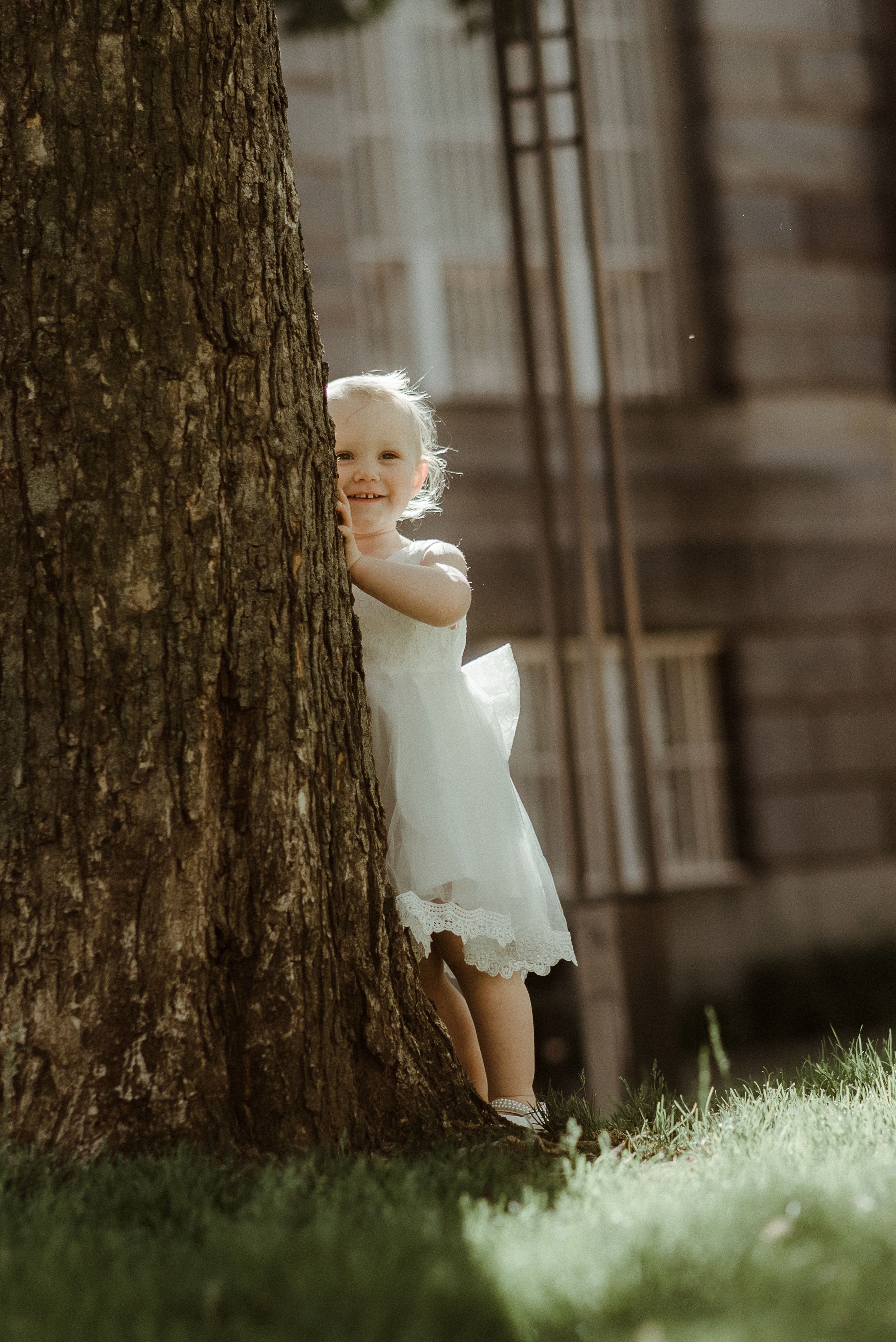 A white dress-clad child peeks playfully from behind a large tree trunk in warm sunlight.