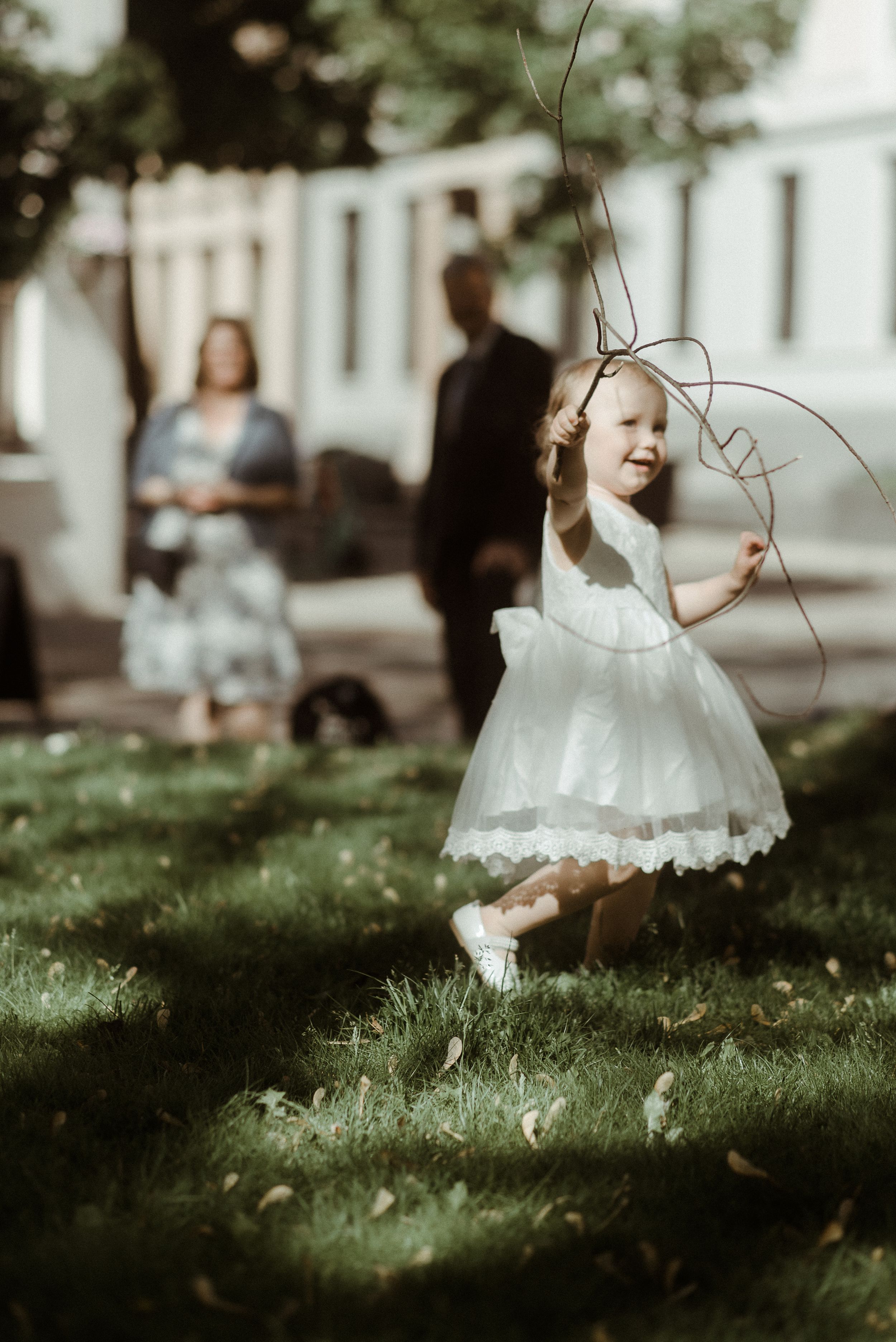 A small child in a white dress twirls in a sunlit garden while adults stand in background.