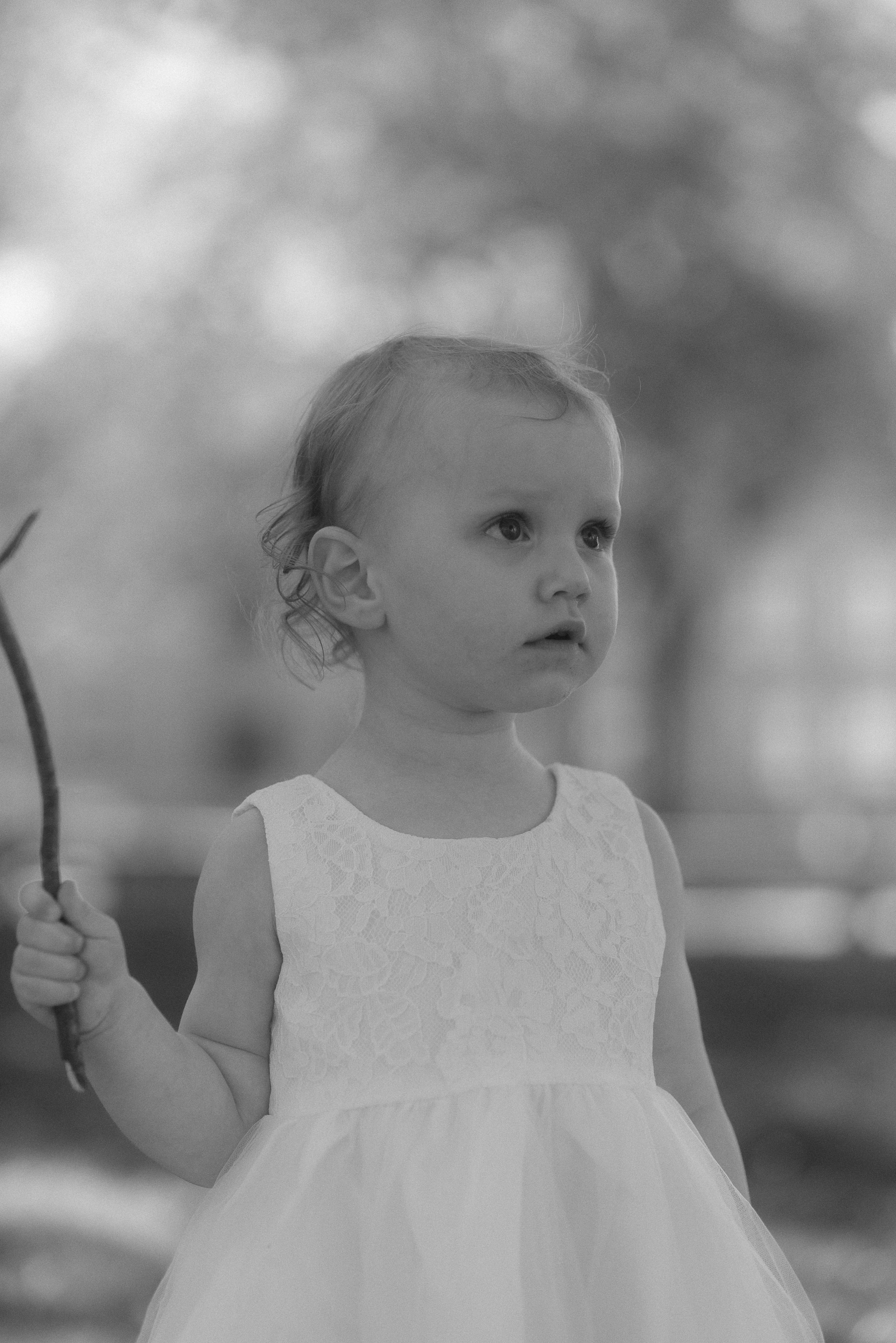 A young child in white dress holds a stick while looking up in black and white photograph.